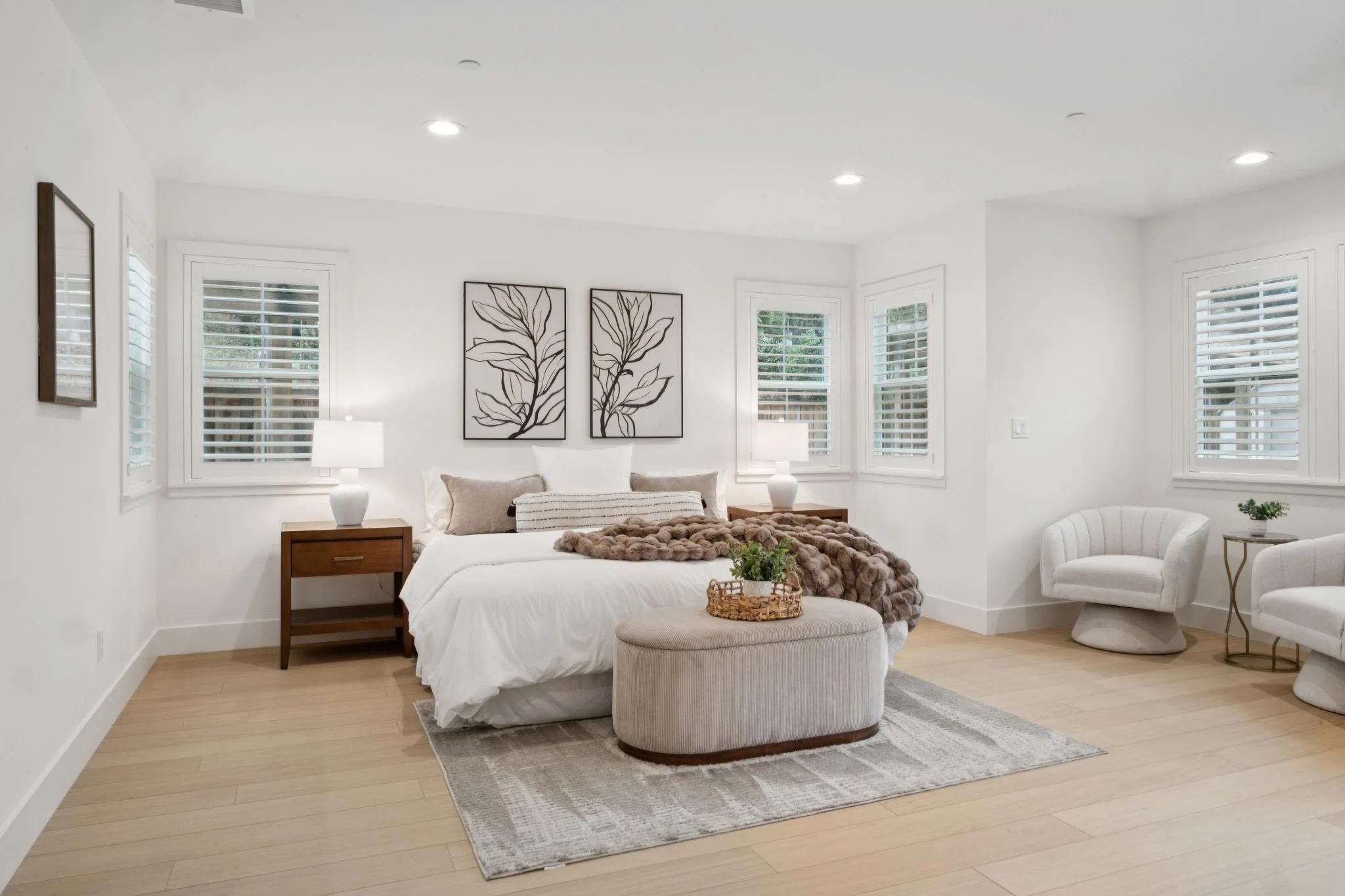 Bright living room with white walls, multiple windows with white shutters, a bed with white bedding and beige pillows, a fluffy brown throw, two white lamps on side tables, black-and-white leaf artwork on the wall, beige ottoman, white armchair, smal