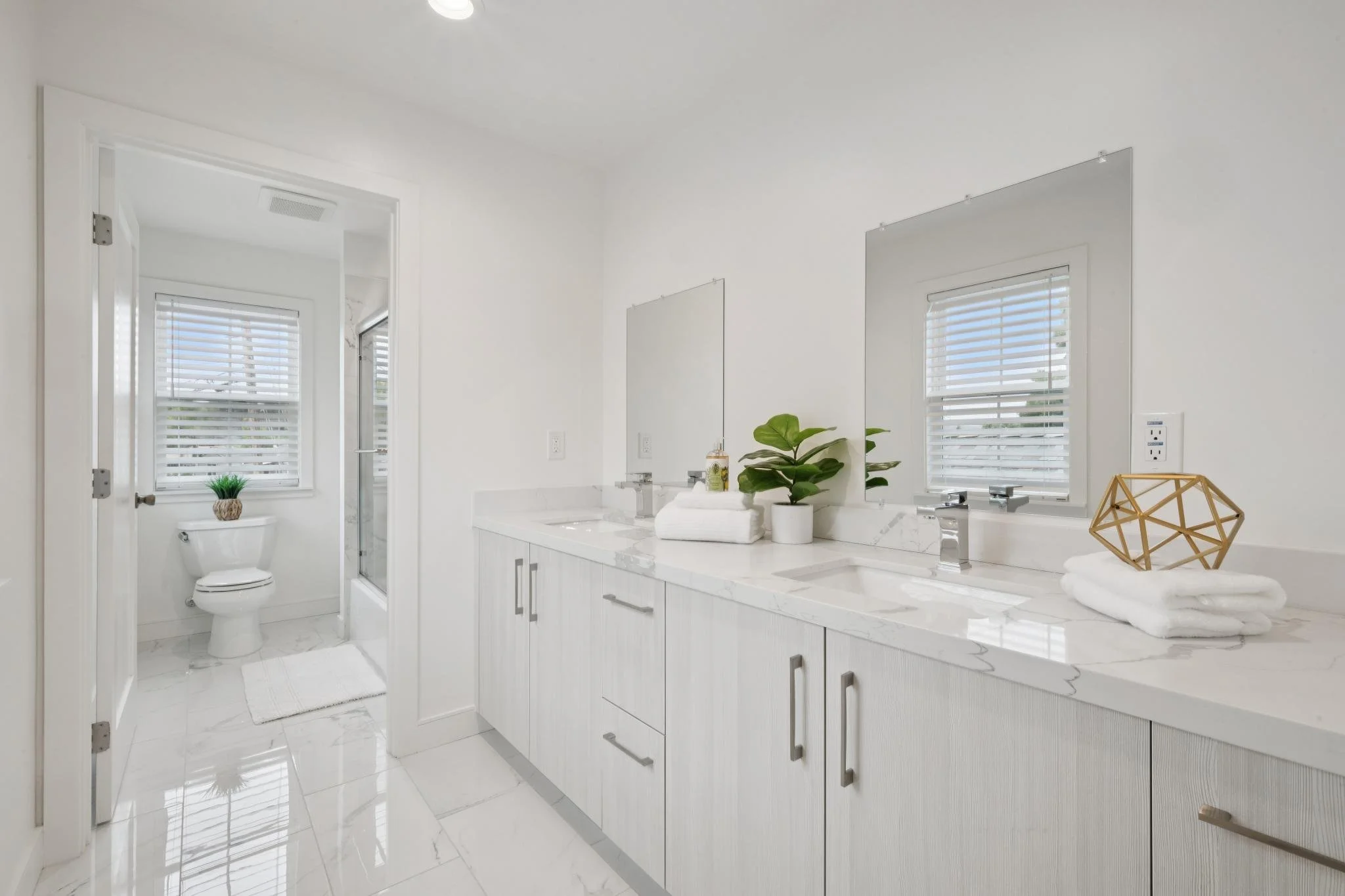 A clean, modern bathroom with white walls, double sinks, and large mirrors. There are two windows with white blinds, a small plant on the toilet tank, and neatly folded towels on the countertop. The floor and countertop are made of white marble, and 