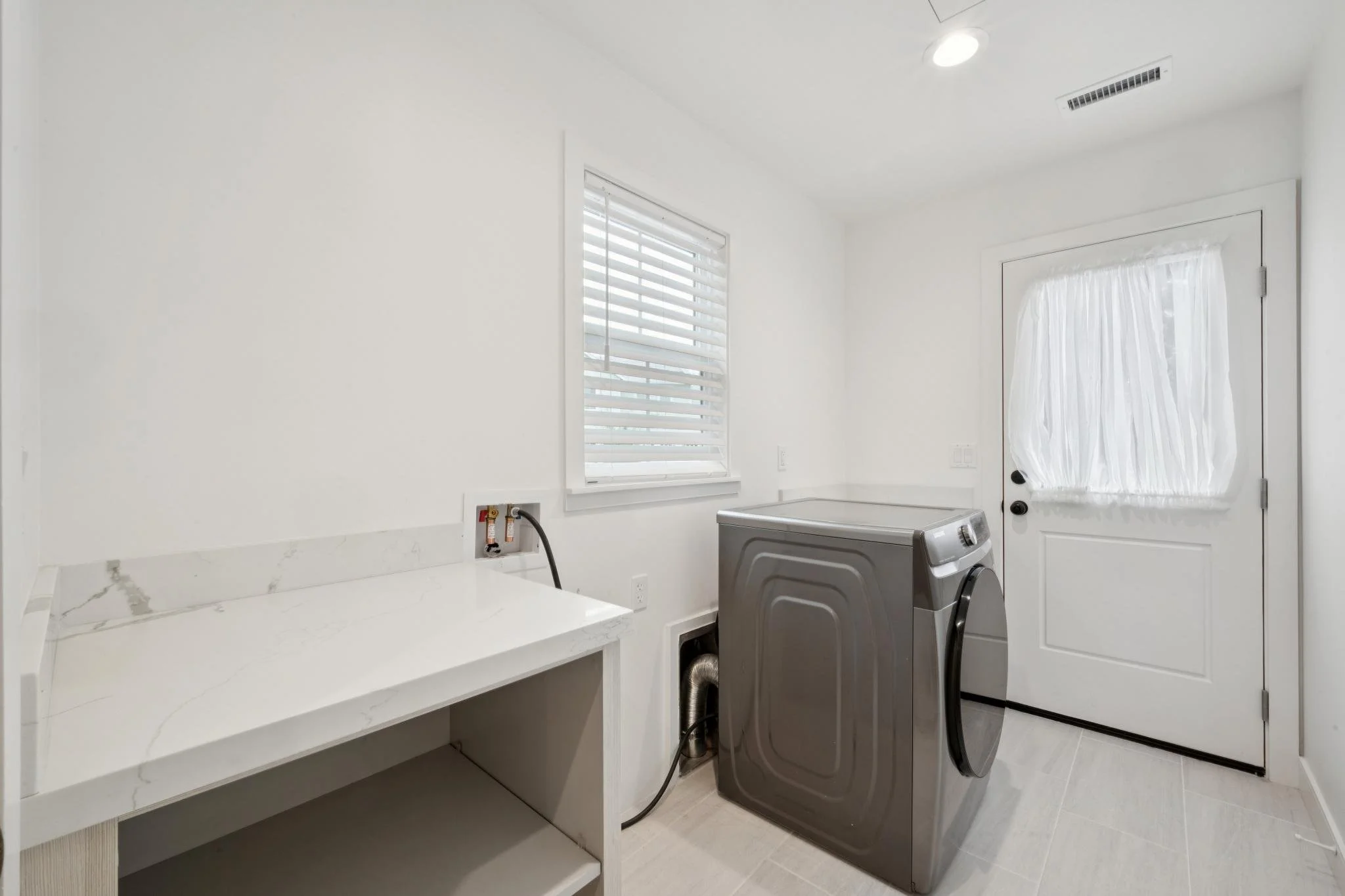 A laundry room with white walls, a window with blinds, a door with sheer curtain, a silver washing machine, white marble countertop, and a white shelf below the counter.