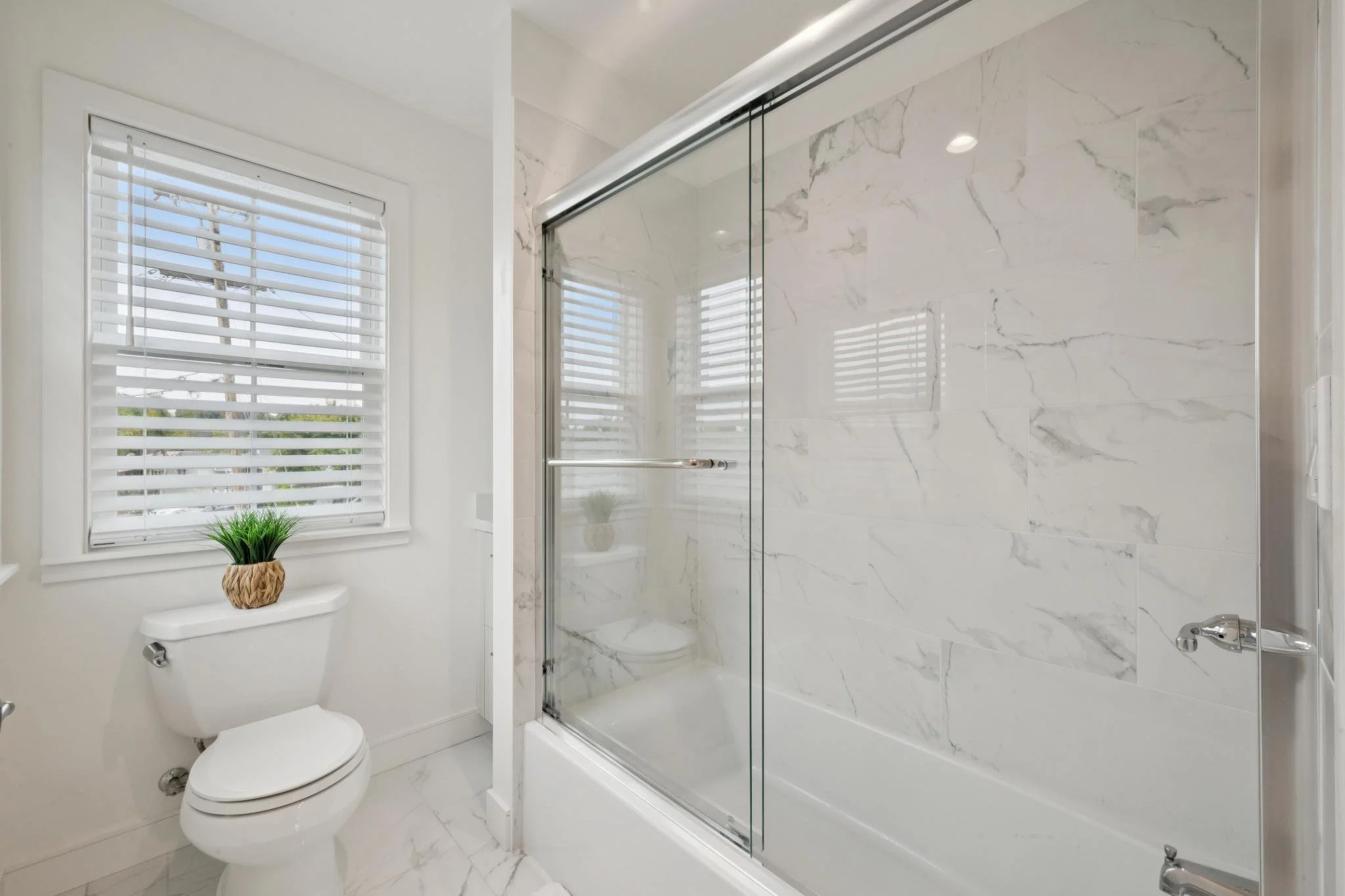 Modern bathroom with a white toilet, a window with white blinds, a potted plant on top of the toilet tank, and a glass-enclosed shower with marble tiles.