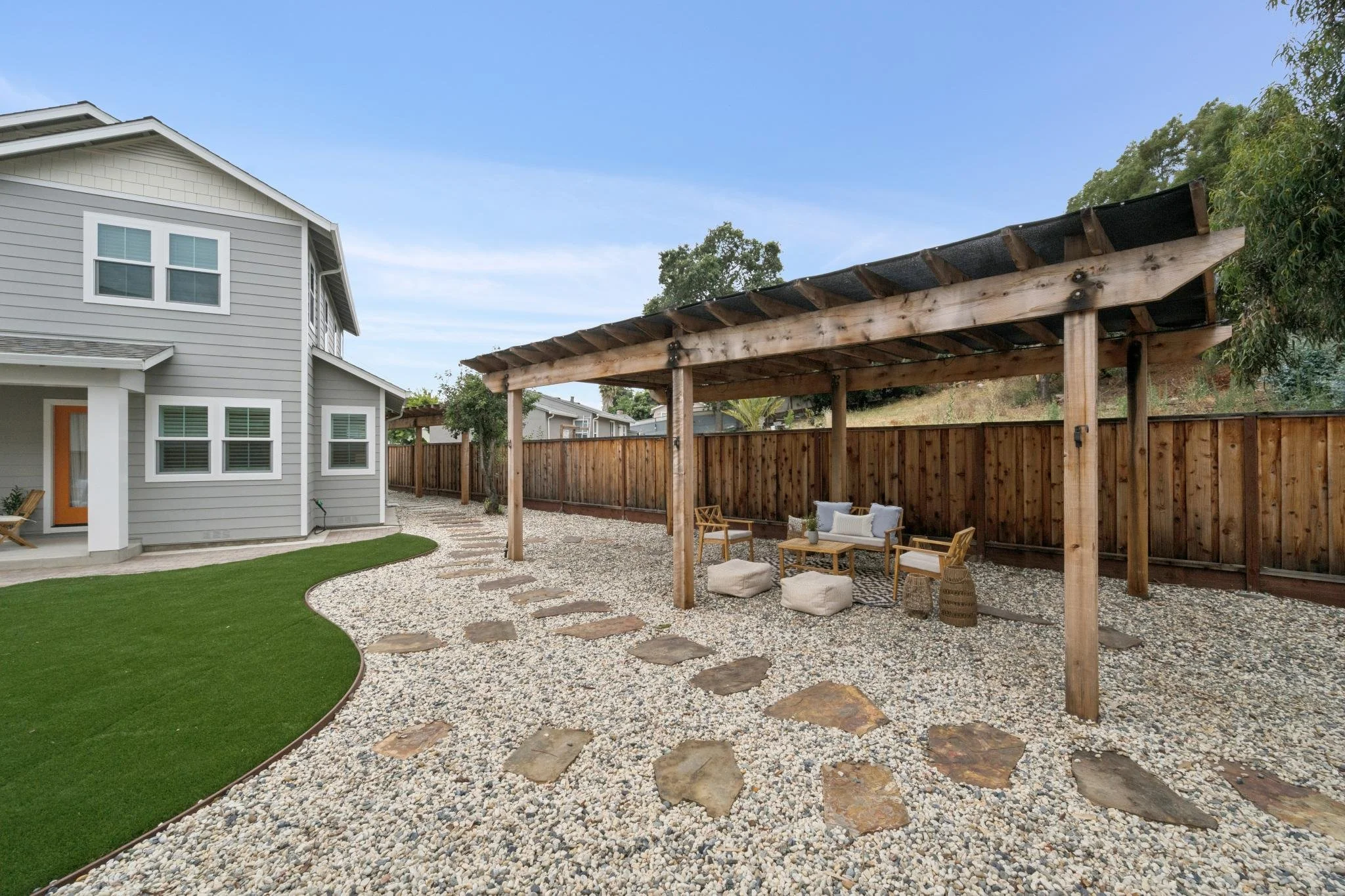 A backyard with a grey two-story house, a wooden pergola with outdoor seating, a pebble and stone pathway, and a fence with trees in the background under a partly cloudy sky.