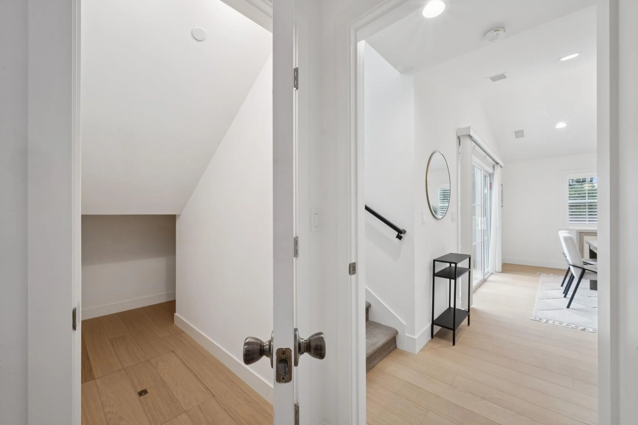 View of a bright, modern interior space with white walls, wooden floors, a staircase, a round mirror on the wall, and a small black table.