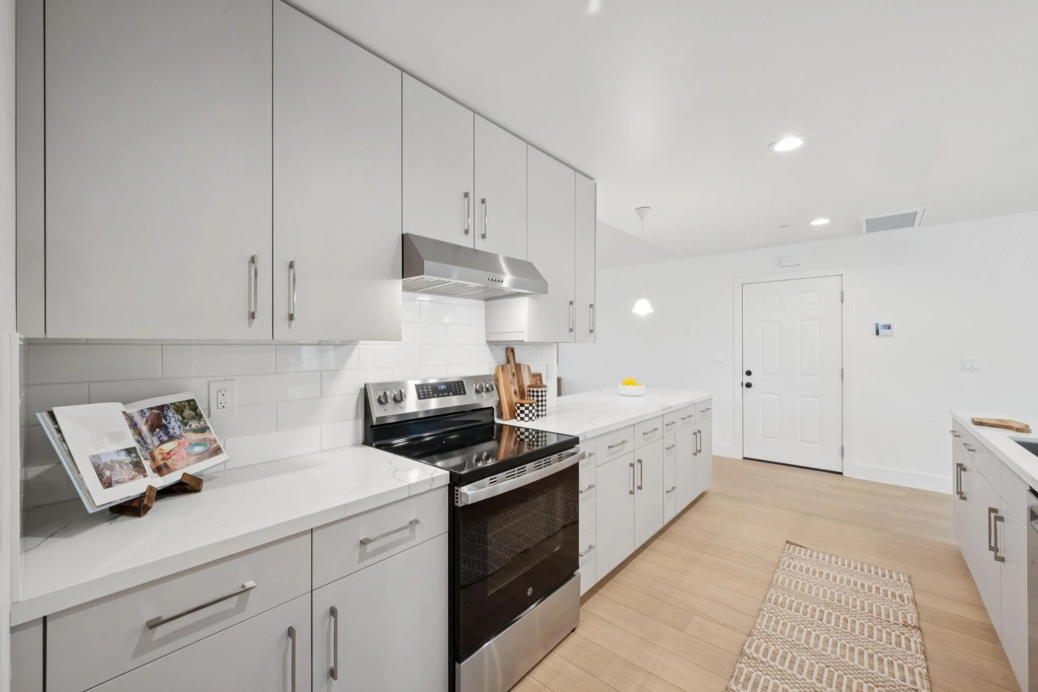 Modern white kitchen with stainless steel appliances, white cabinets, light wood flooring, and a small rug in front of the door, featuring a bowl of lemons on the countertop.