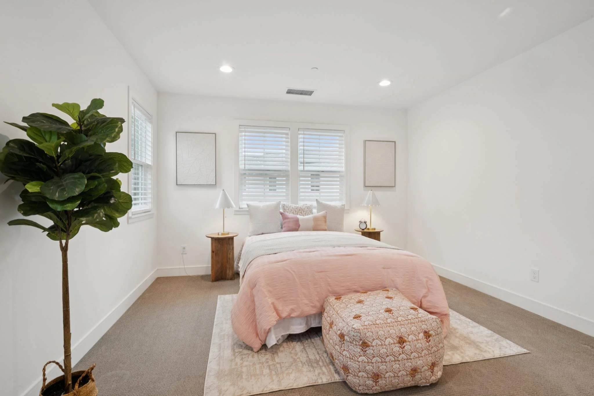 A bedroom with a bed covered in a pink bedspread, a patterned ottoman at the foot of the bed, two white lamps on wooden nightstands, and framed artwork on the walls. There is a green potted plant on the left side, white window blinds behind the bed, 