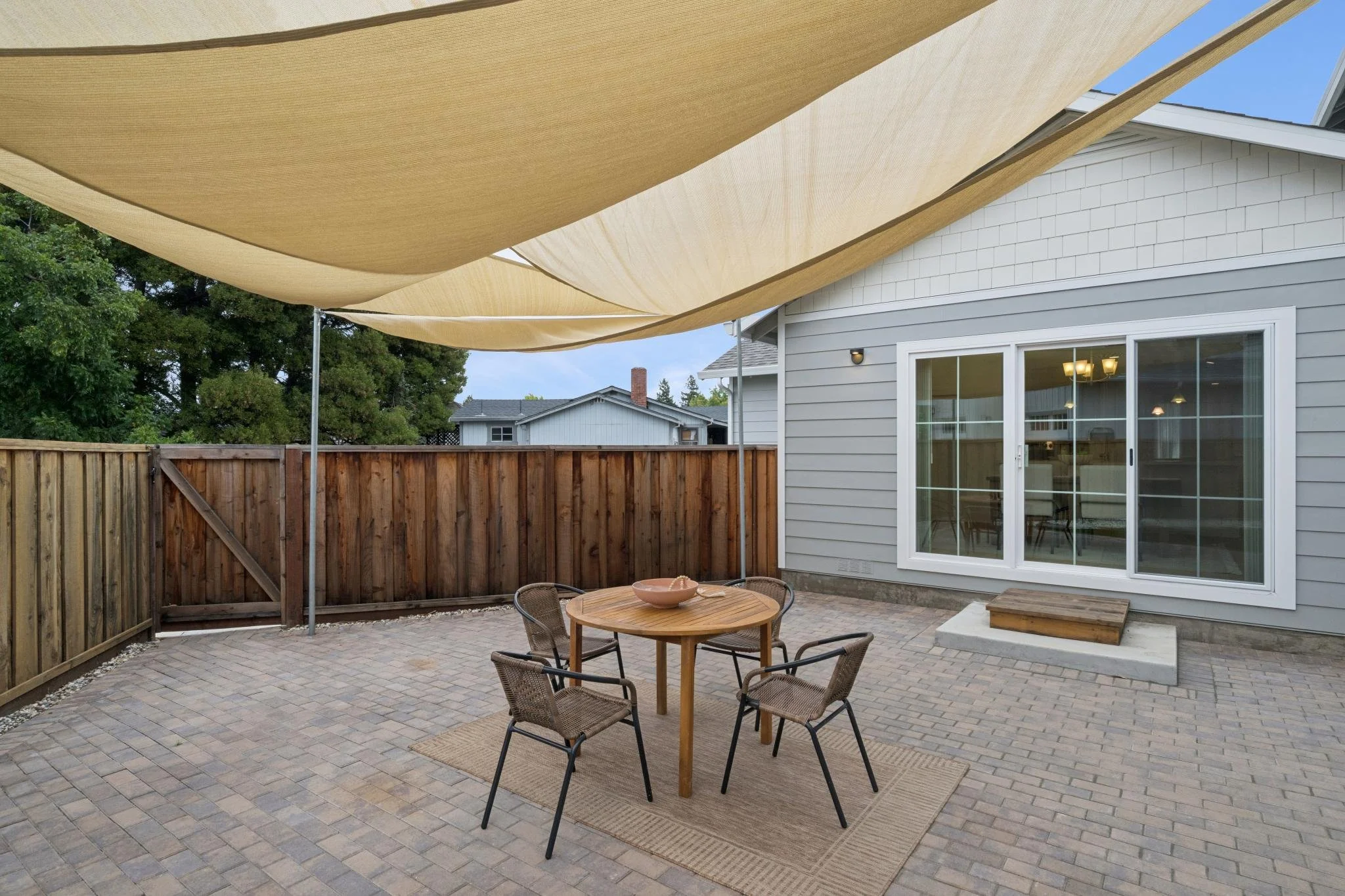 Residential backyard patio with a table and four chairs, a rug, a beige sunshade sail, a wooden fence, and a house with sliding glass door and exterior light