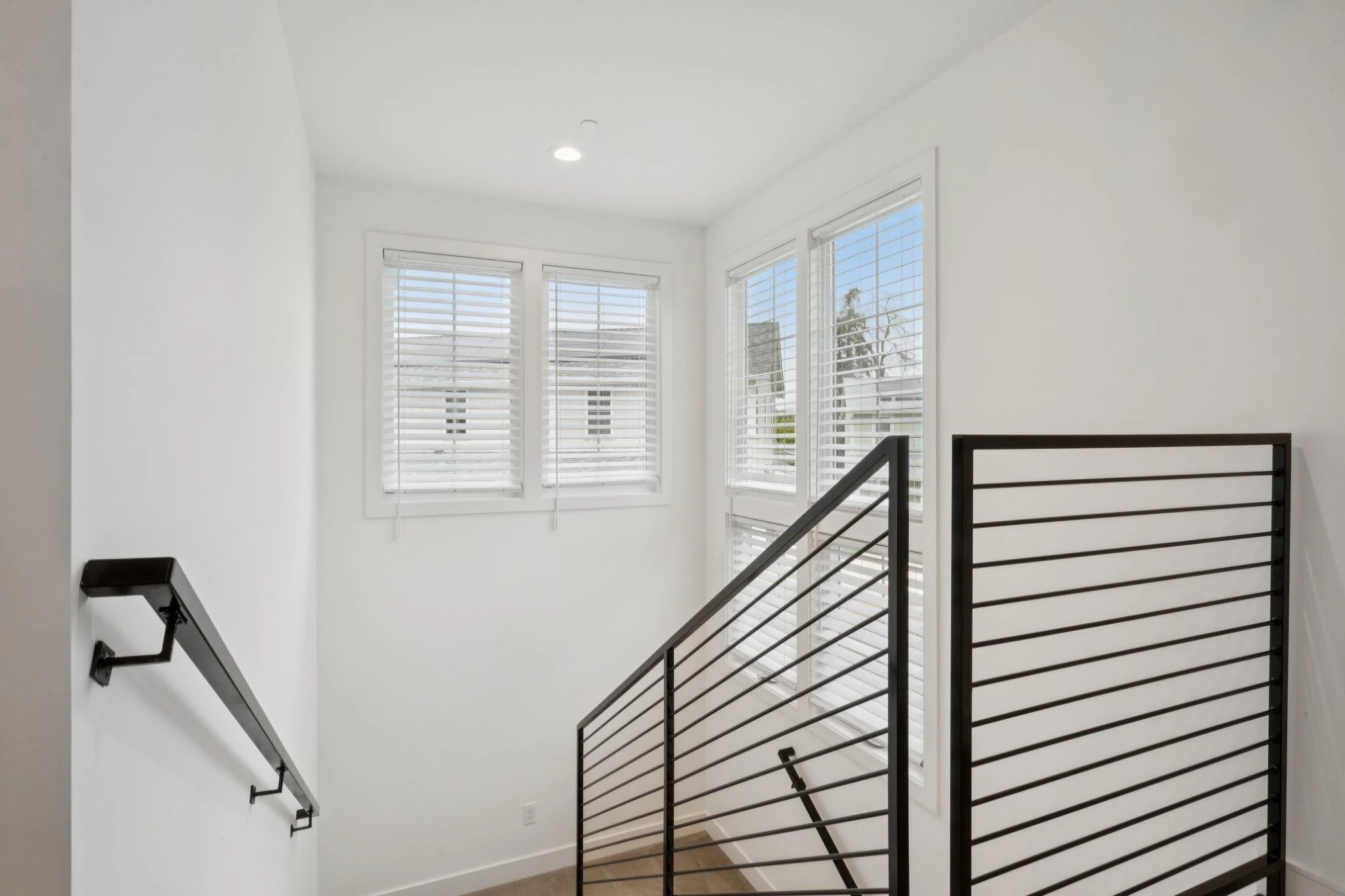 Interior view of a stairwell with white walls, large windows with blinds, a black metal railing, and ceiling lights.