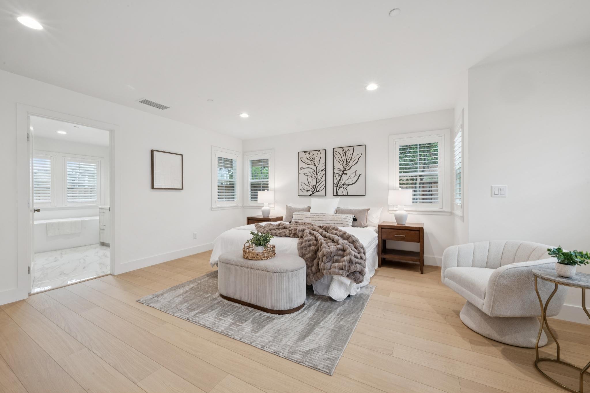 Bright bedroom with white walls and large windows. A bed with a fur blanket, pillows, and a small ottoman at the foot. Nightstands with lamps on each side. Framed artwork on the wall behind the bed. Light-colored hardwood floors and a rug under the b