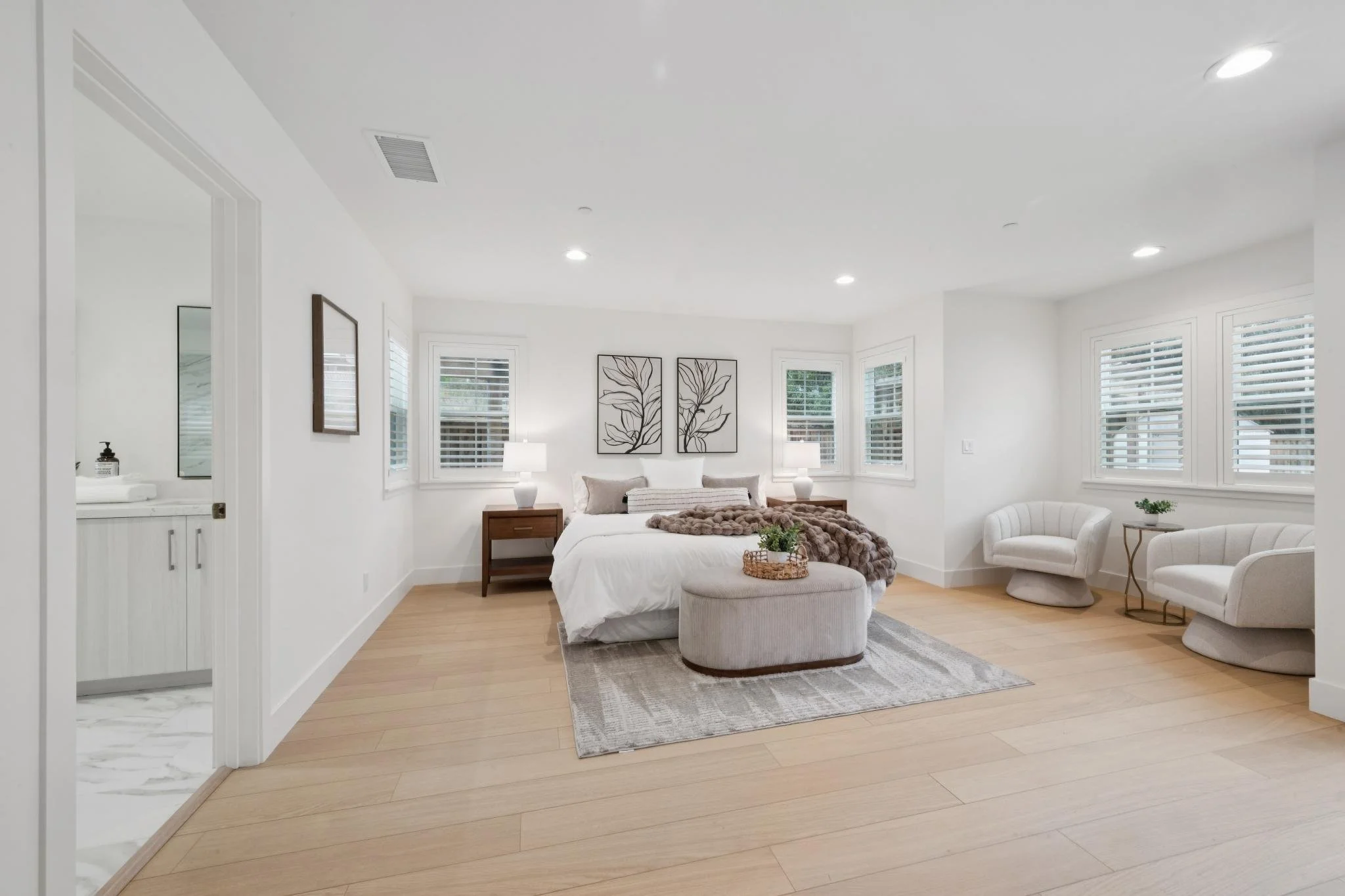 Bright bedroom with white walls, hardwood floors, a bed with white bedding, two wooden nightstands with lamps, and framed botanical art on the wall. Contains window blinds, a small cream-colored armchair, and a patterned gray and white area rug.