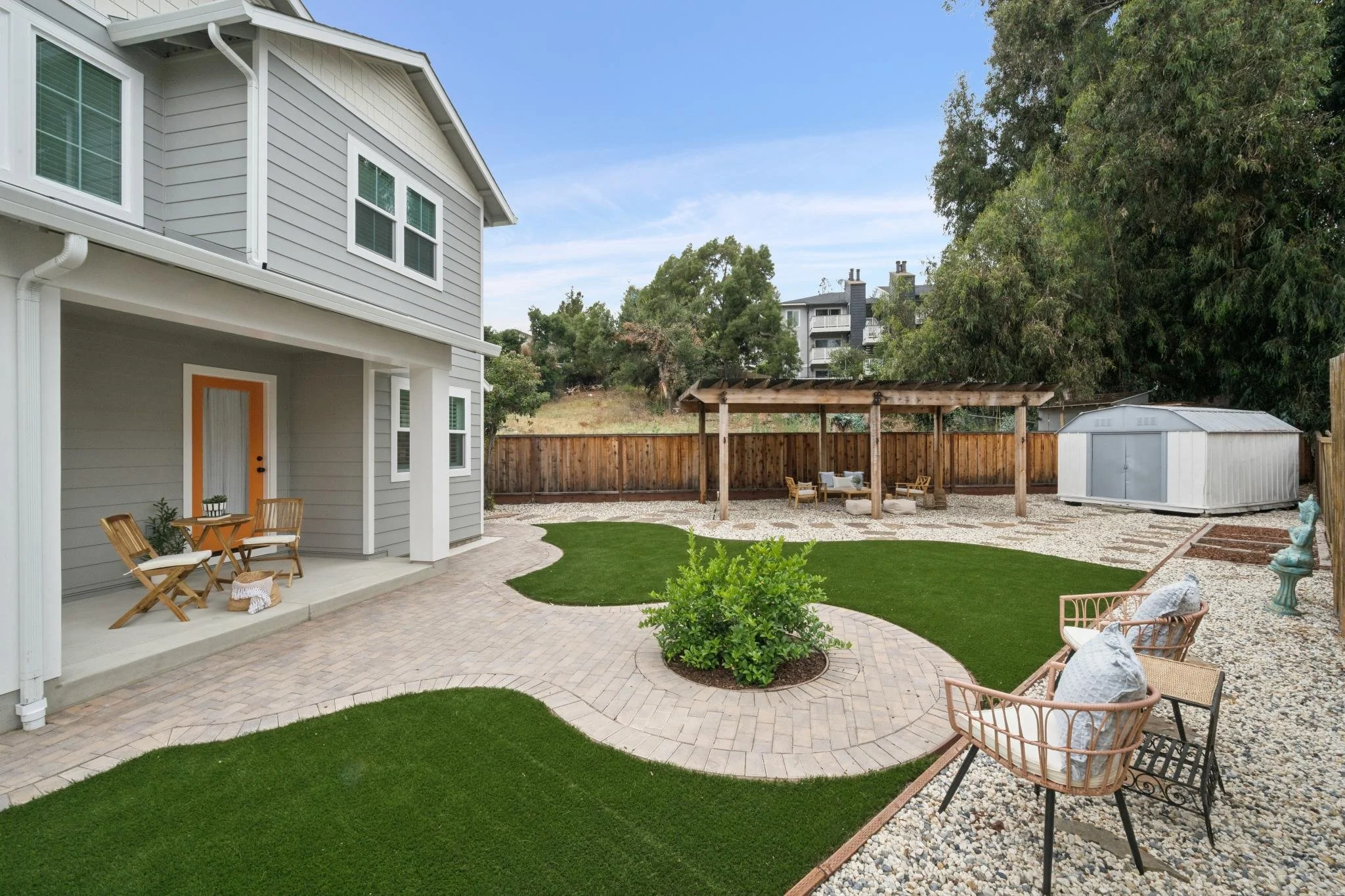 A backyard with a patio, a small round table with four chairs, a green lawn, a gravel area with outdoor seating under a wooden pergola, a large green tree, a white shed, and a wooden fence, with houses on a hillside in the background.