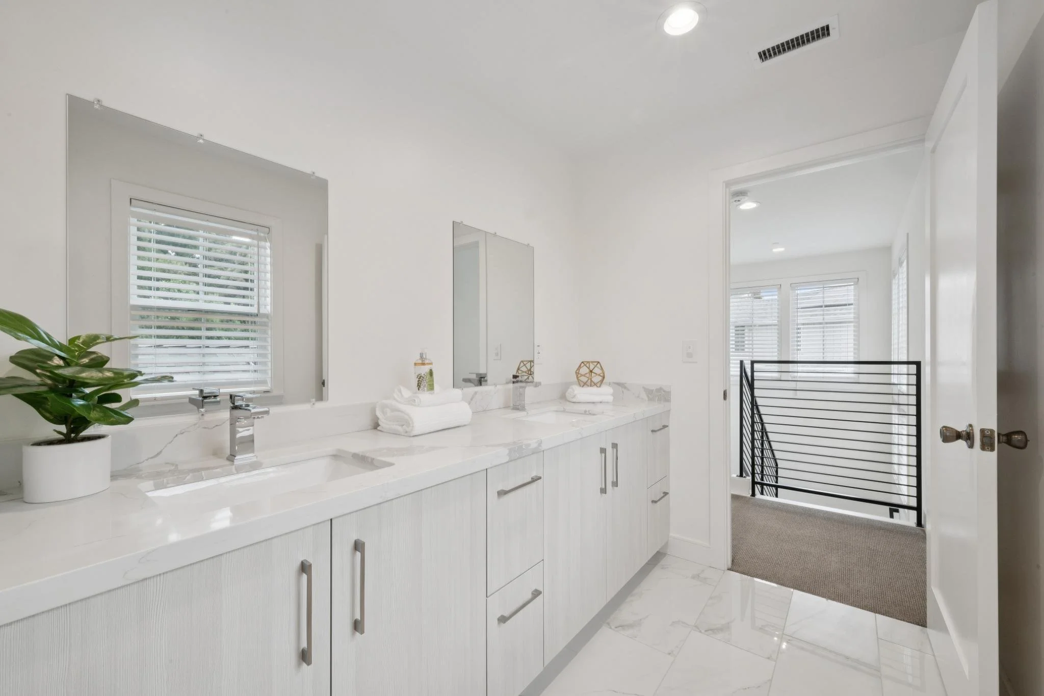 Modern bathroom with white cabinetry, marble countertop, large mirror, windows with blinds, green potted plant, and towel sets, with adjoining hallway.