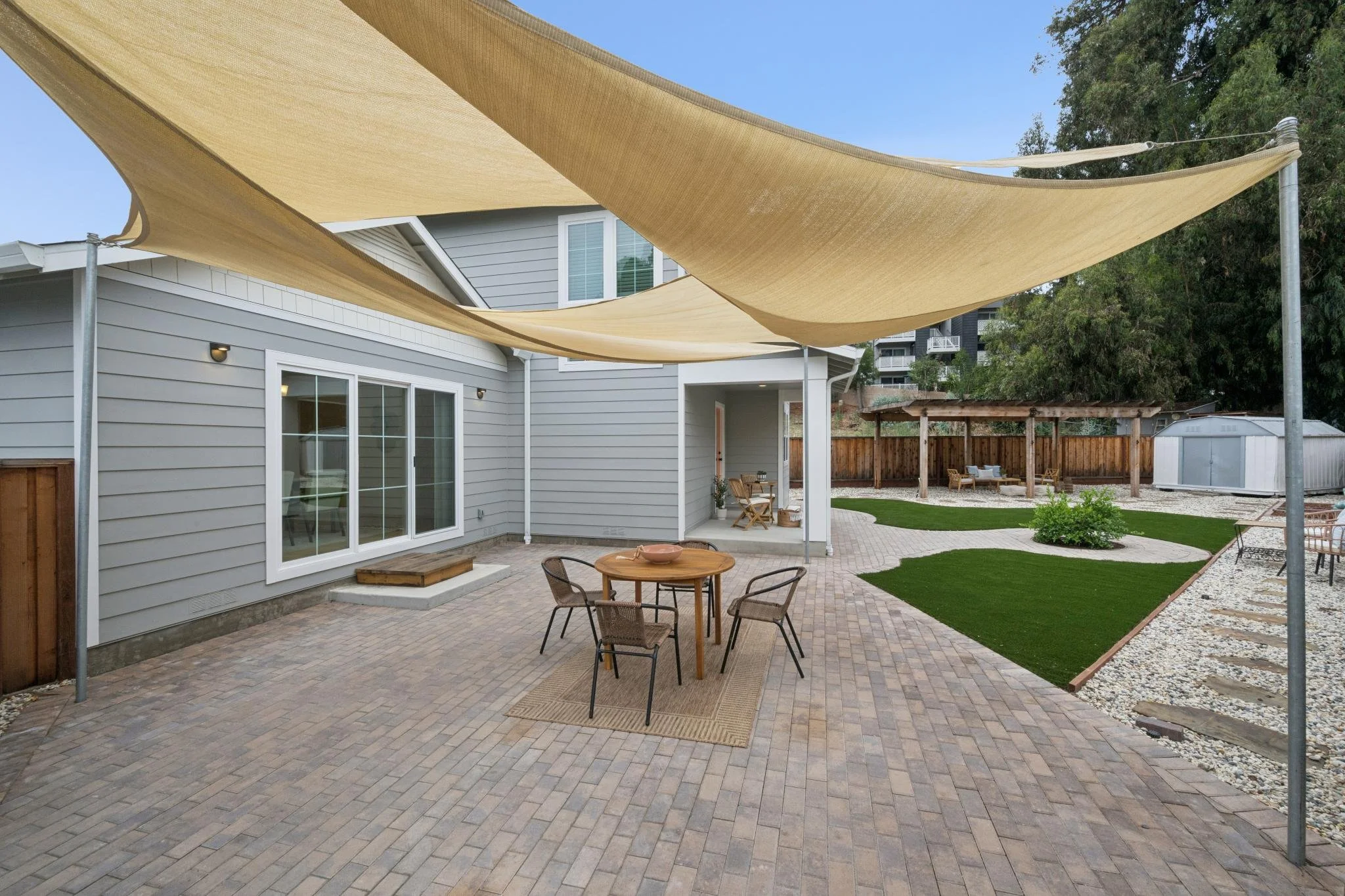 Backyard patio with beige shade sails, wooden table, chairs, a small garden area with grass and bushes, and a house with large windows and sliding door.