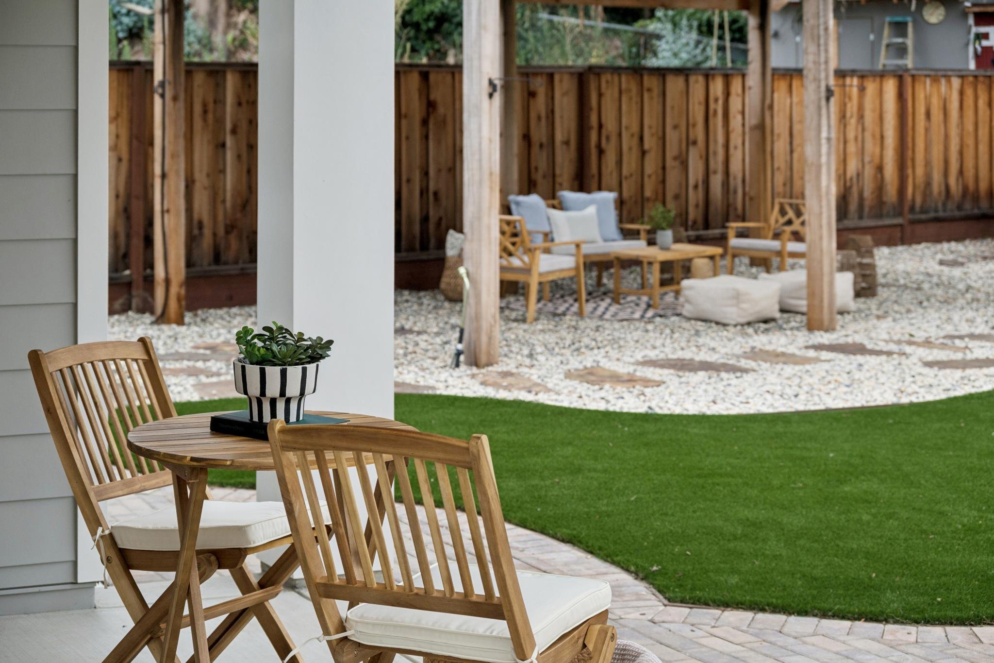 Backyard patio with wooden chairs, a table with a plant, and a fenced area with outdoor seating and a rug.