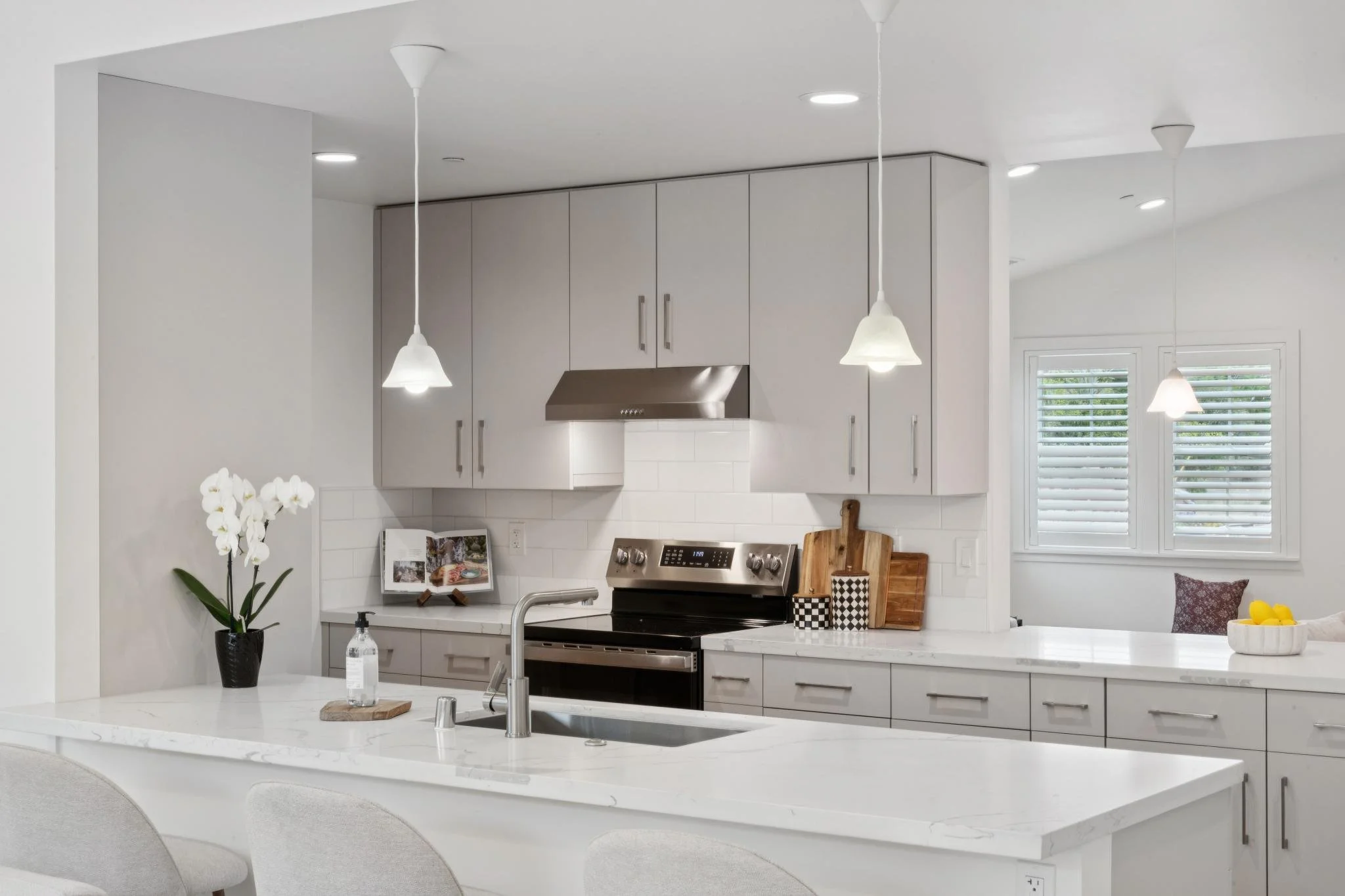 Modern kitchen with white island, gray cabinets, stainless steel stove, white countertops, and window with white shutters.
