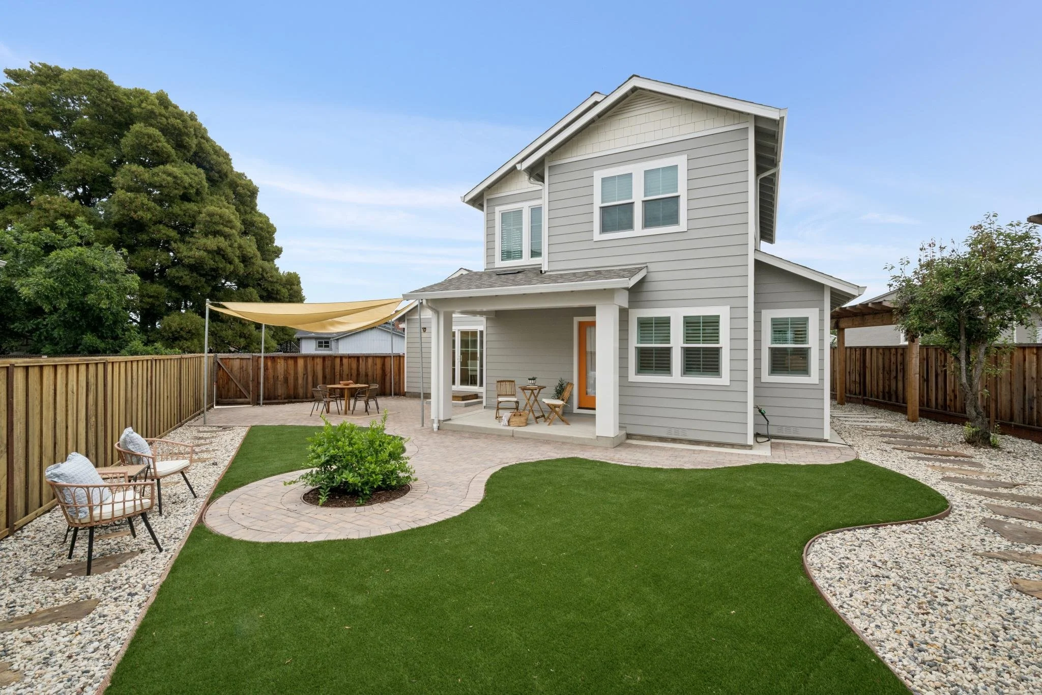 Backyard with a modern two-story house, fenced yard, patio furniture, a sunshade, patches of artificial grass, and a garden with trees.