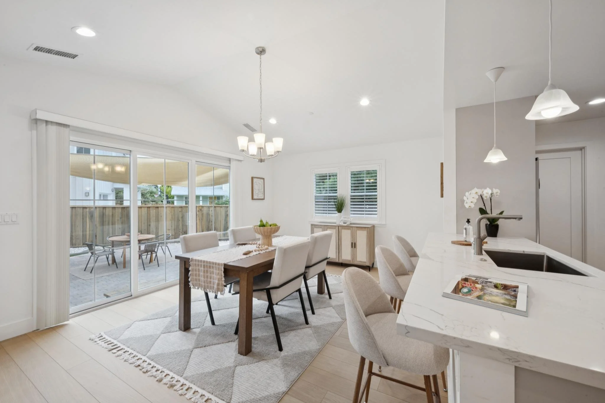 A modern, bright dining and kitchen area with large sliding glass doors leading to an outdoor patio. The dining table has a beige and white checkered table runner and cream-colored chairs. Pendant lights hang from the ceiling, and there's a white mar