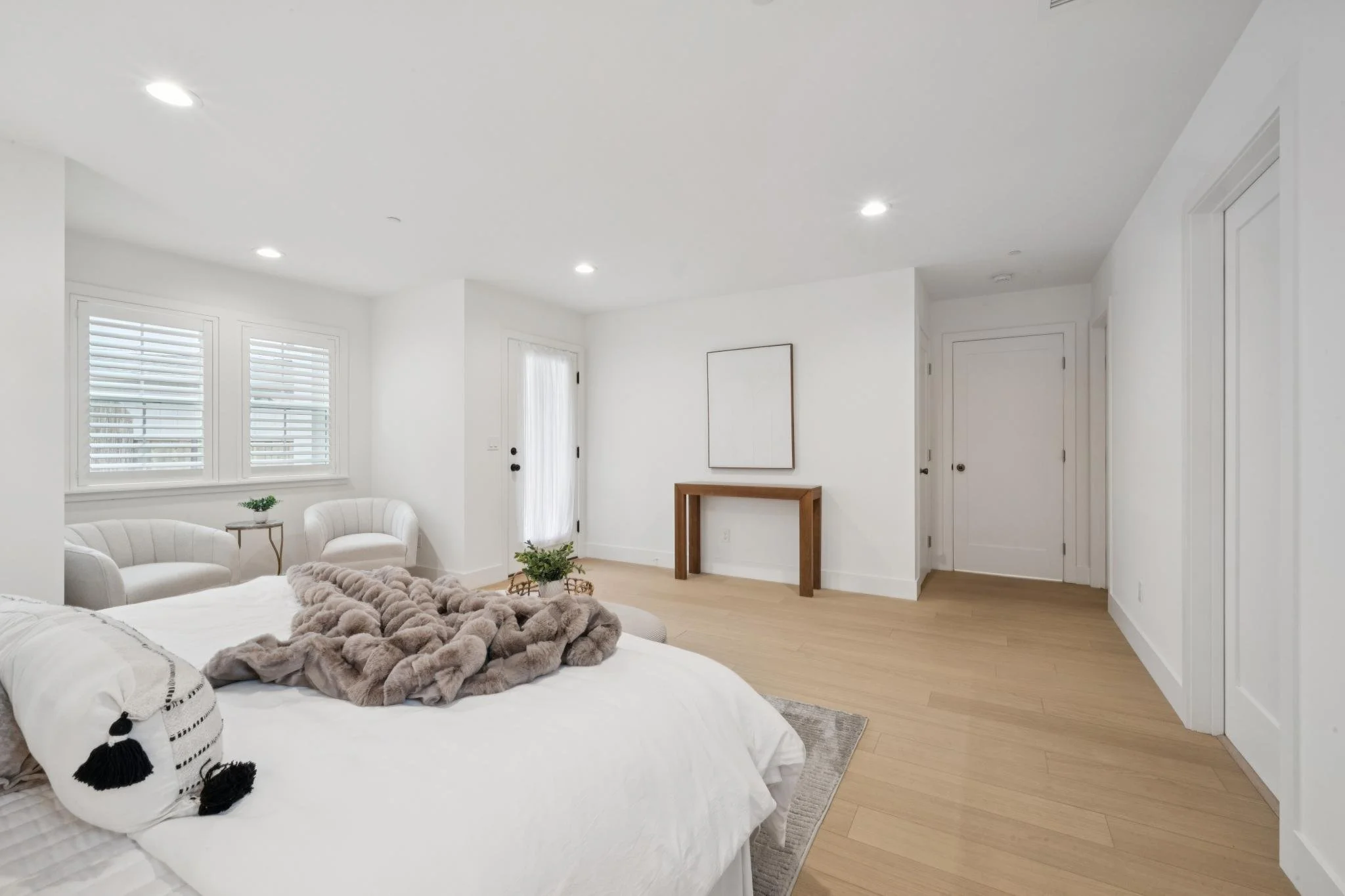 A bright, modern living room with white walls, two windows with white blinds, a white sofa with black and white tasseled pillows, a plush blanket, a wooden side table with a plant, and a minimalist wooden console table against the wall.