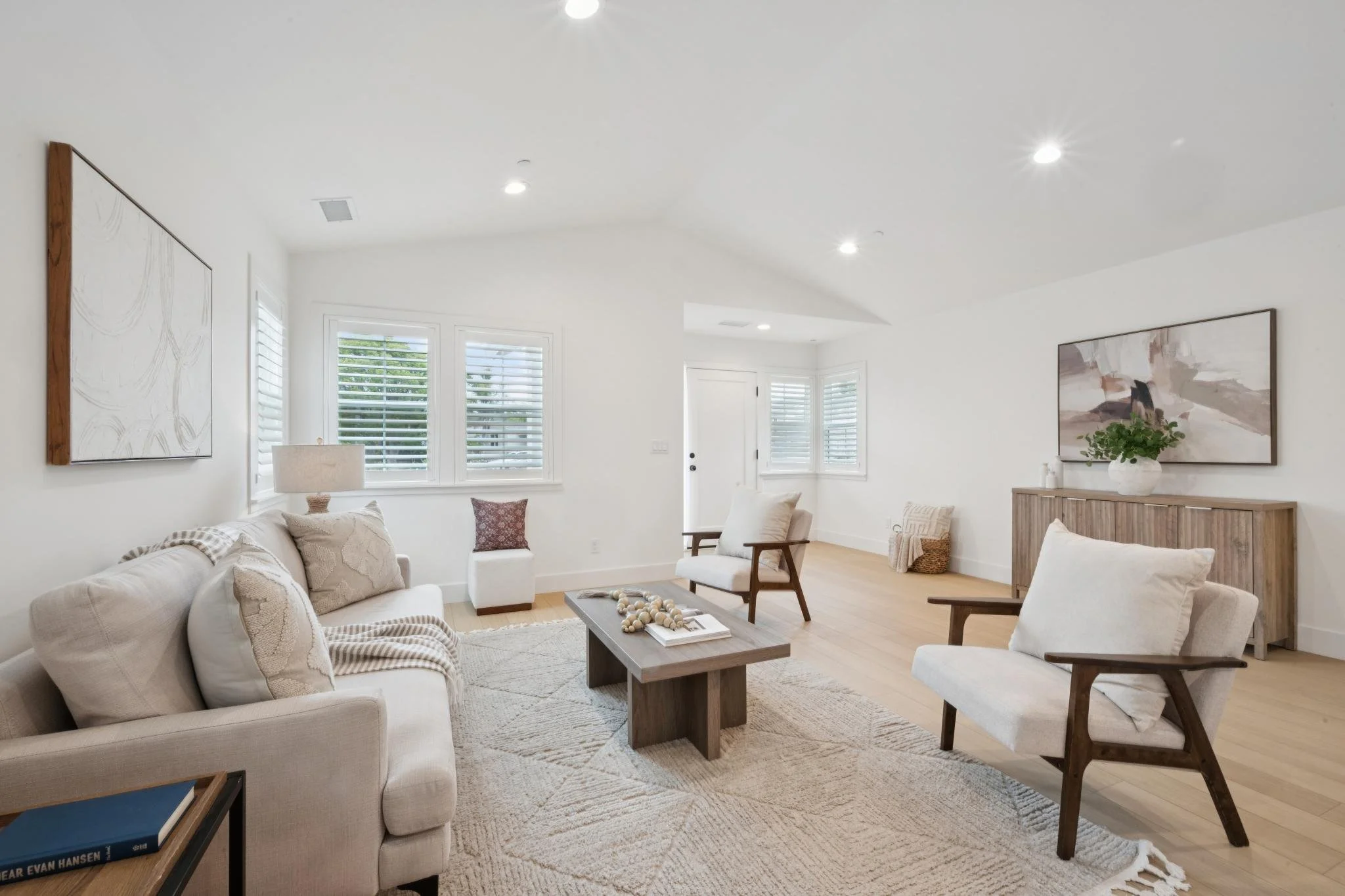 Bright living room with white walls, multiple windows with shutters, and modern furniture including a beige sofa, wooden chairs, and a coffee table with decor. Artwork and a potted plant adorn the walls and surface.