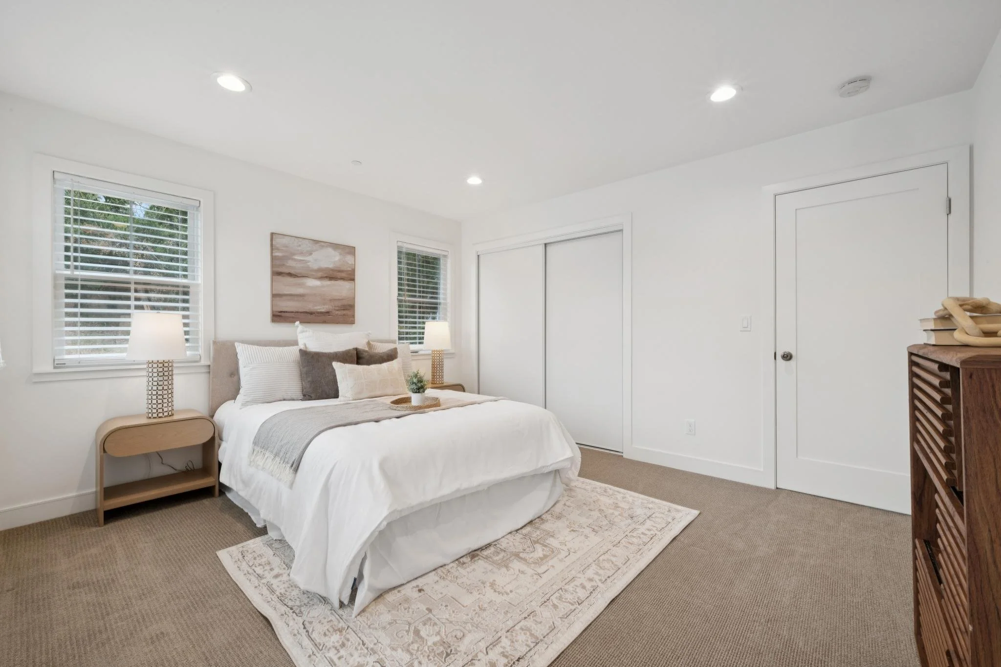 A bedroom with a bed, bedside tables, lamps, windows with blinds, a picture on the wall, a closet with sliding doors, a wooden dresser, and a rug on a beige carpet.