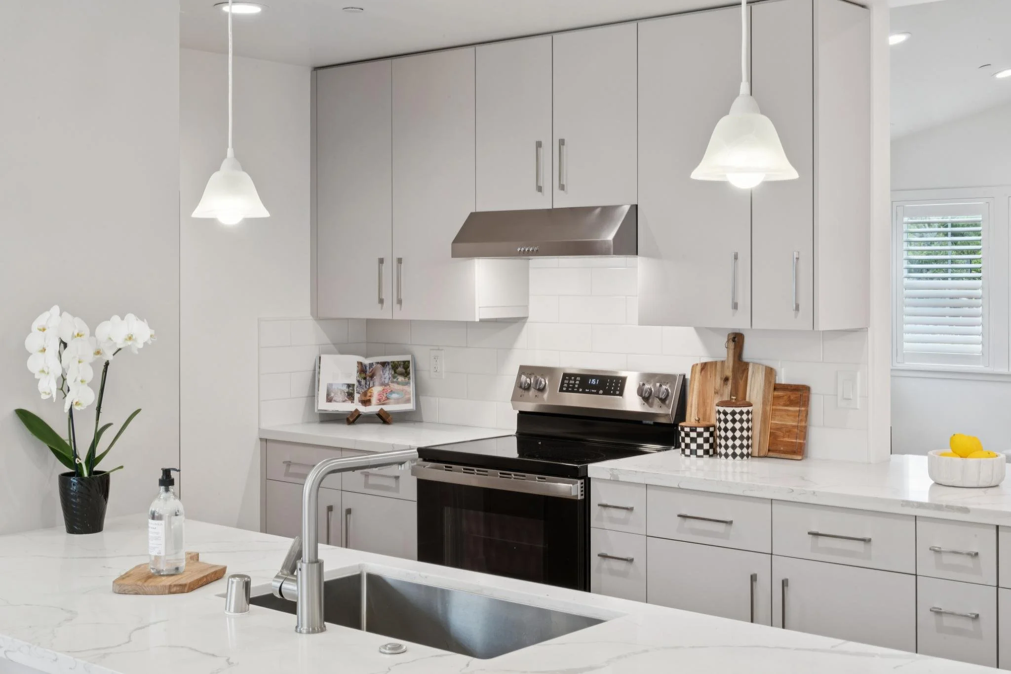 A modern kitchen with white cabinets, a stainless steel range hood, black stove, white subway tile backsplash, white marble countertops, and a window with plantation shutters. The kitchen island has a white flower vase with orchids, a soap dispenser,