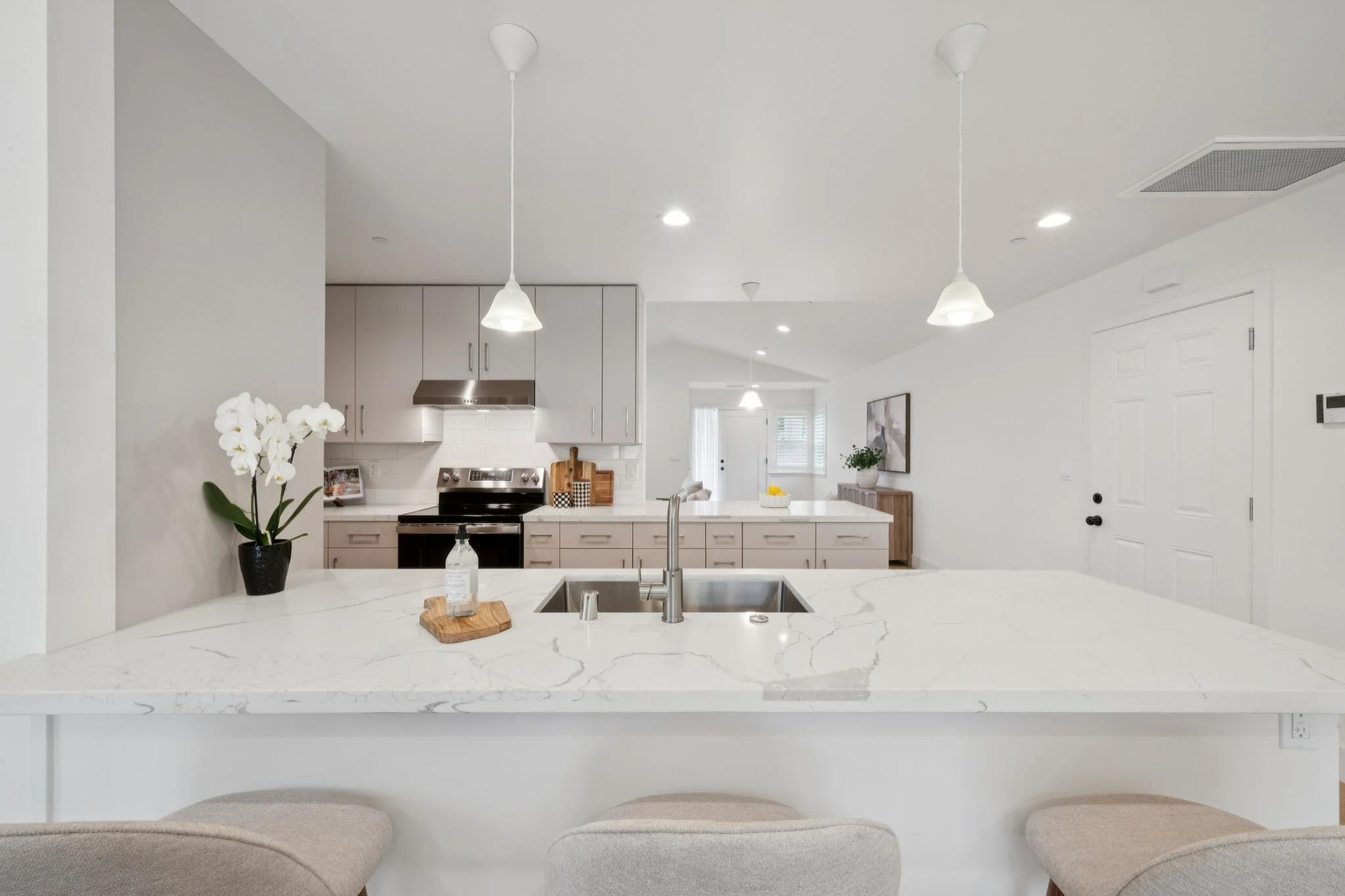 Modern kitchen with white countertops, light gray cabinets, stainless steel stove, pendant lighting, a potted orchid, and a kitchen island with a sink and chairs.