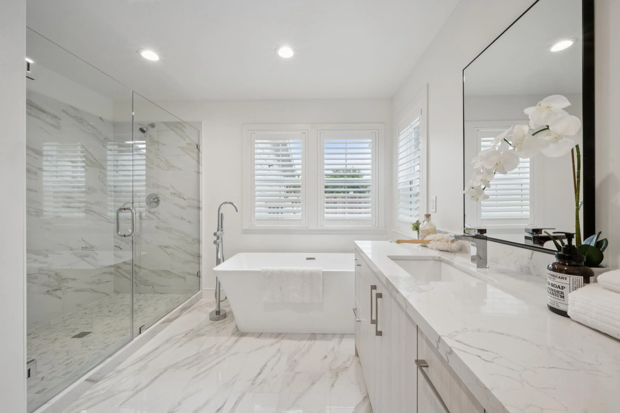 Modern white bathroom with large window, bathtub, and walk-in shower with glass door, marble tile surfaces, and a mirror with flowers on the vanity.