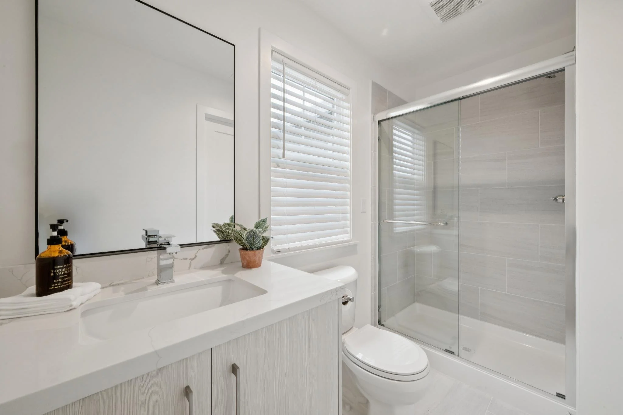 A modern bathroom with a white marble vanity, a rectangular mirror, and a potted plant. There are two amber bottles on the counter, towels underneath, and a glass-enclosed shower with beige tiles. A window with white blinds is above the sink, letting