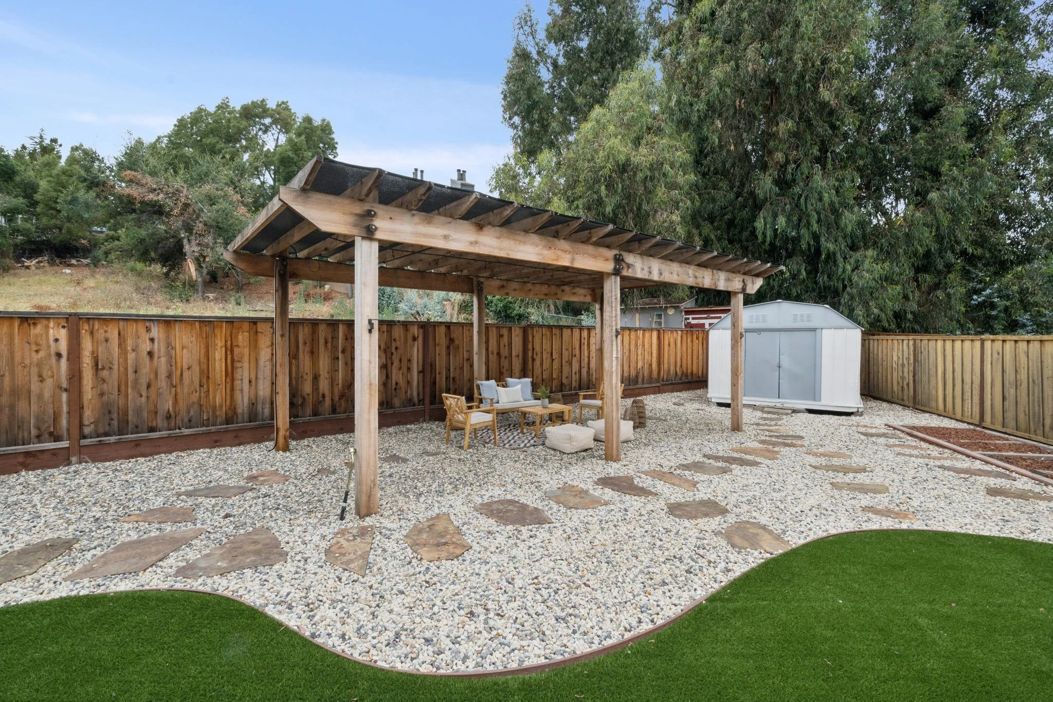 A backyard with a wooden pergola, outdoor furniture, a gravel ground cover with stepping stones, and a tall wooden fence. There are trees and a white shed in the background.