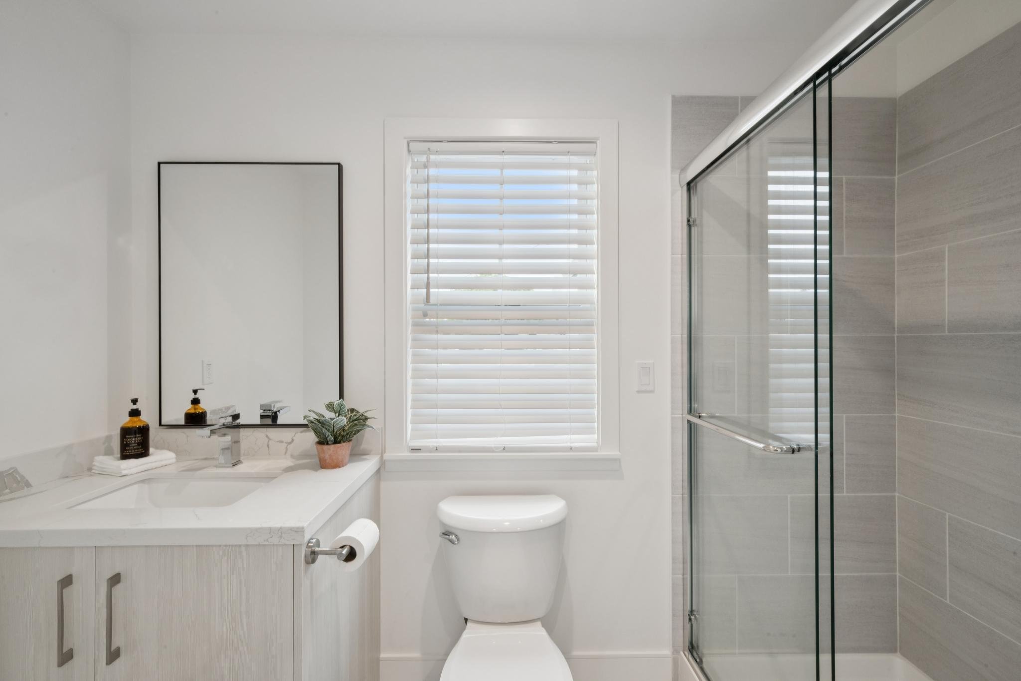 A modern bathroom with a white vanity, a rectangular mirror, a potted plant, soap bottles, a toilet, and a glass shower enclosure with beige tile walls, a window with white blinds, and a white ceiling.