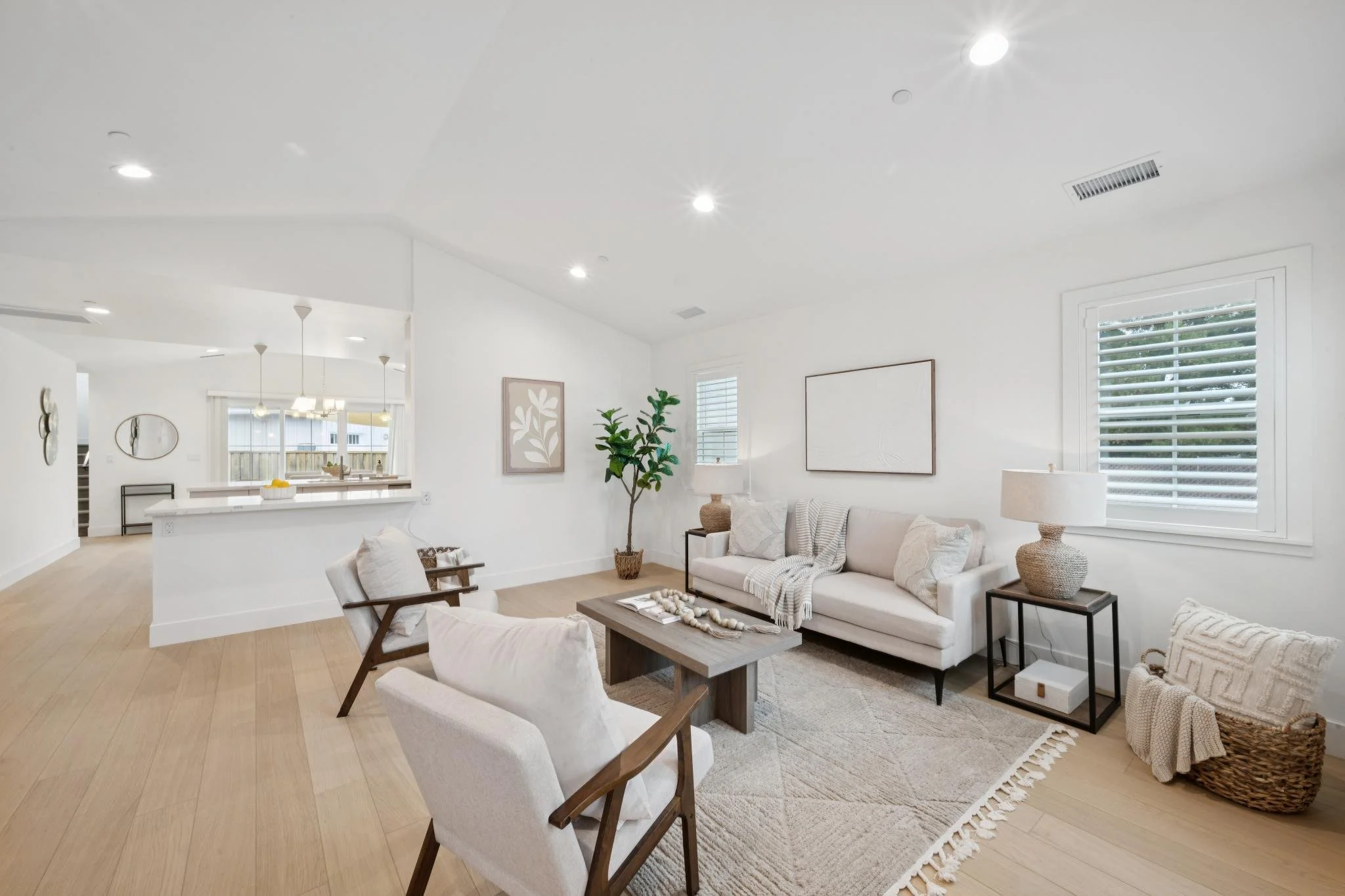 A bright, modern living room with white walls, large windows with white blinds, and contemporary furniture including a white sofa with throw pillows. Decor includes a large potted plant, framed artwork, and a beige table lamp. The space opens to a dining area and kitchen with pendant lights.