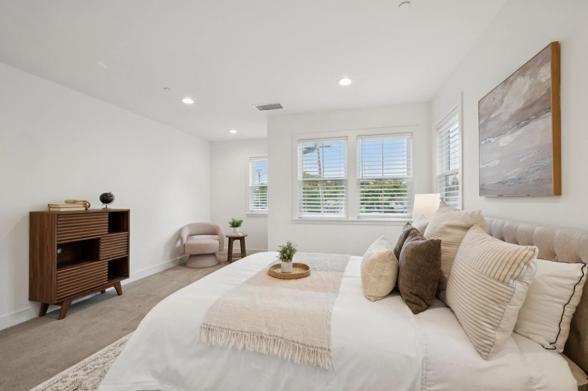 A bright living room with white walls, four windows with blinds, beige carpet, and modern furniture including a beige sofa with multiple pillows, a pink armchair, a wooden side table, and a decorative artwork on the wall.