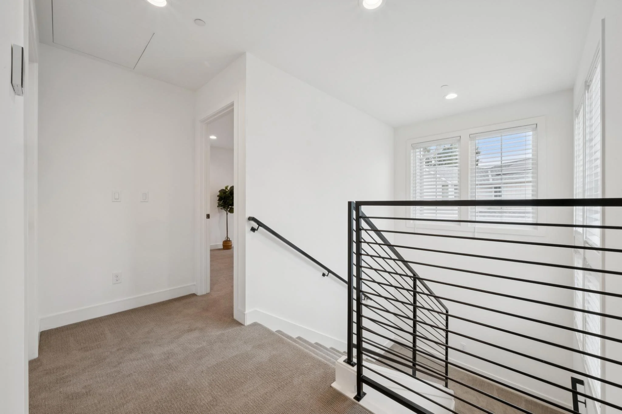 Bright, modern staircase area with white walls, beige carpeted floor, large windows with blinds, and a black metal railing.
