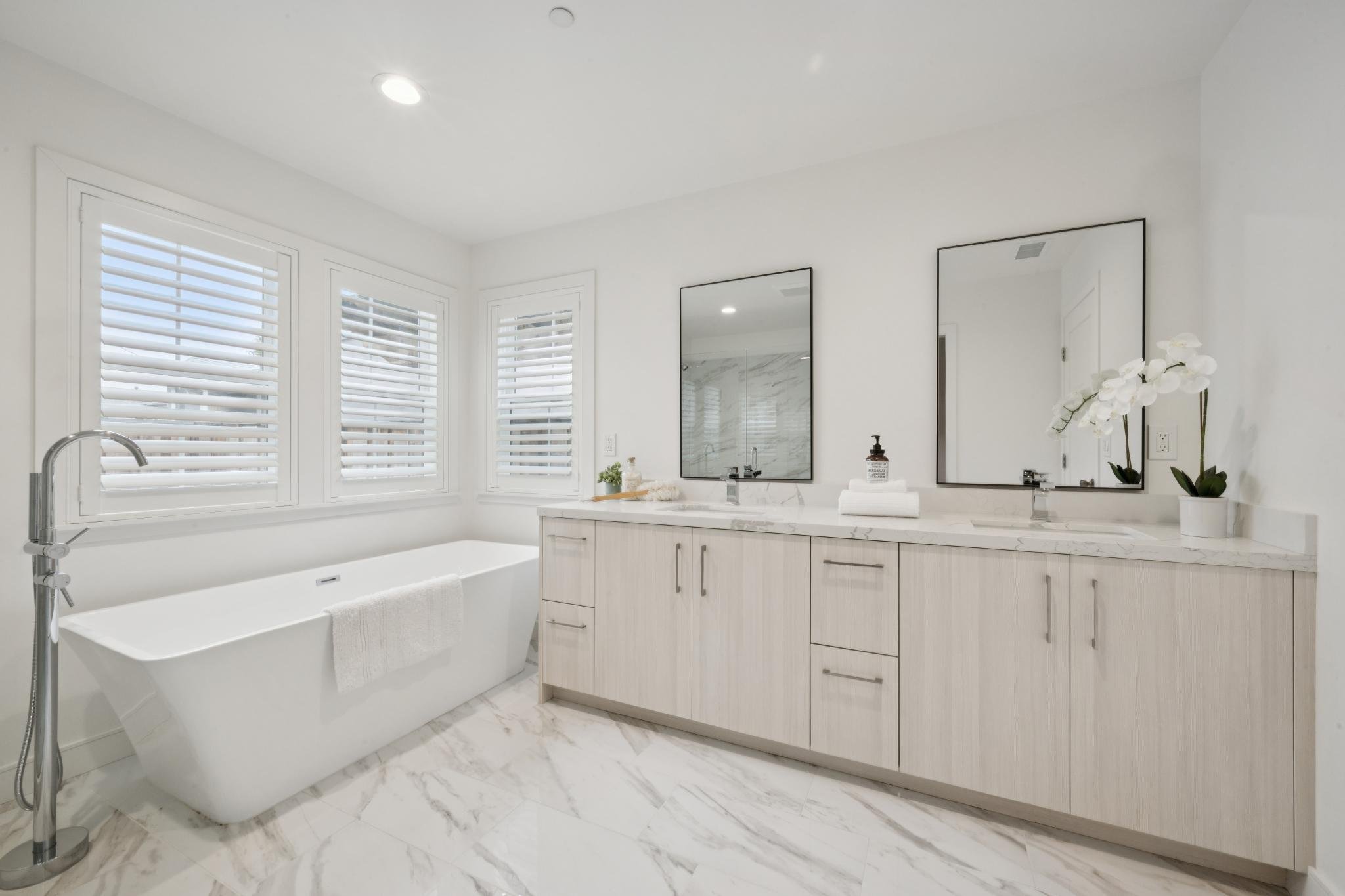 Bright modern bathroom with white marble flooring, white vanity, large mirrors, orchid plant, soap dispenser, and bathtub underneath windows with white shutters.