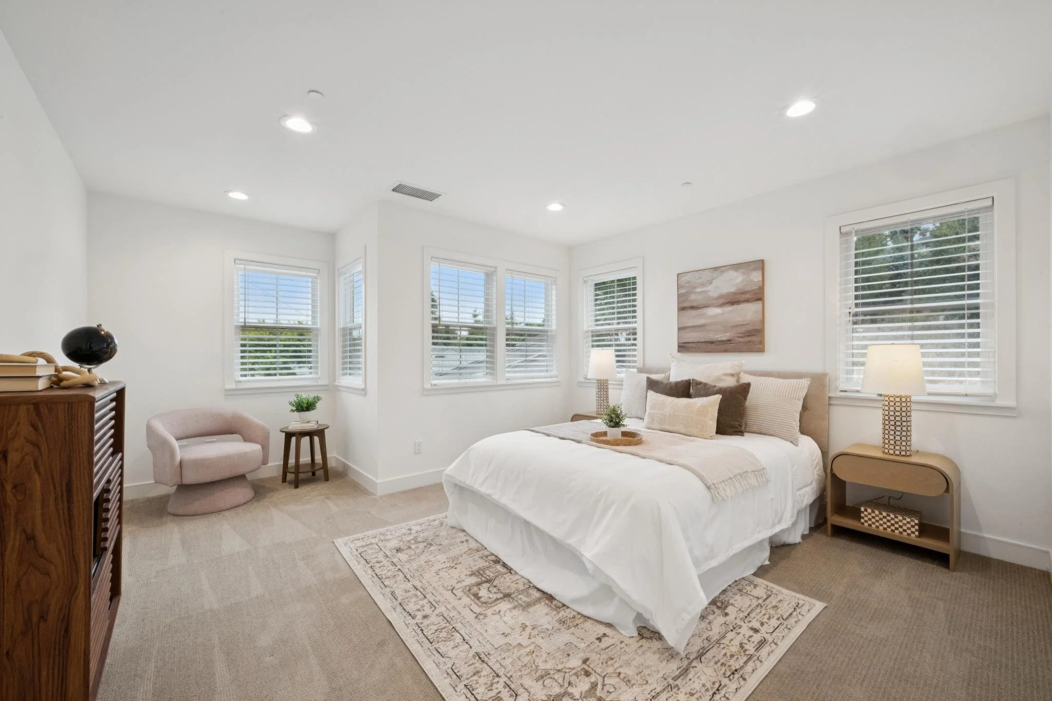 Bright bedroom with large windows, bed with white bedding, beige and brown pillows, wooden side tables, lamps, and a painting on the wall.