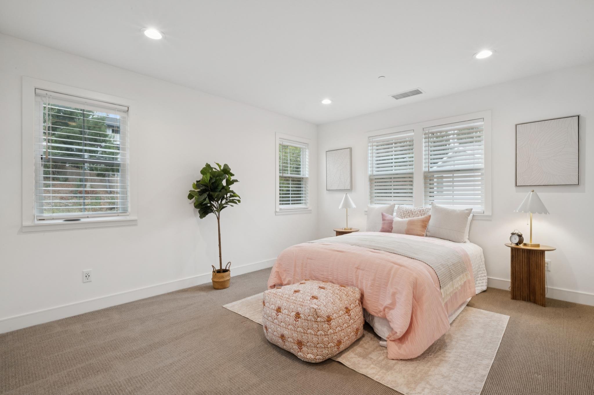 Bright, modern bedroom with white walls, multiple windows with blinds, a bed with light pink bedding, a small round ottoman, a potted plant, and two bedside lamps.