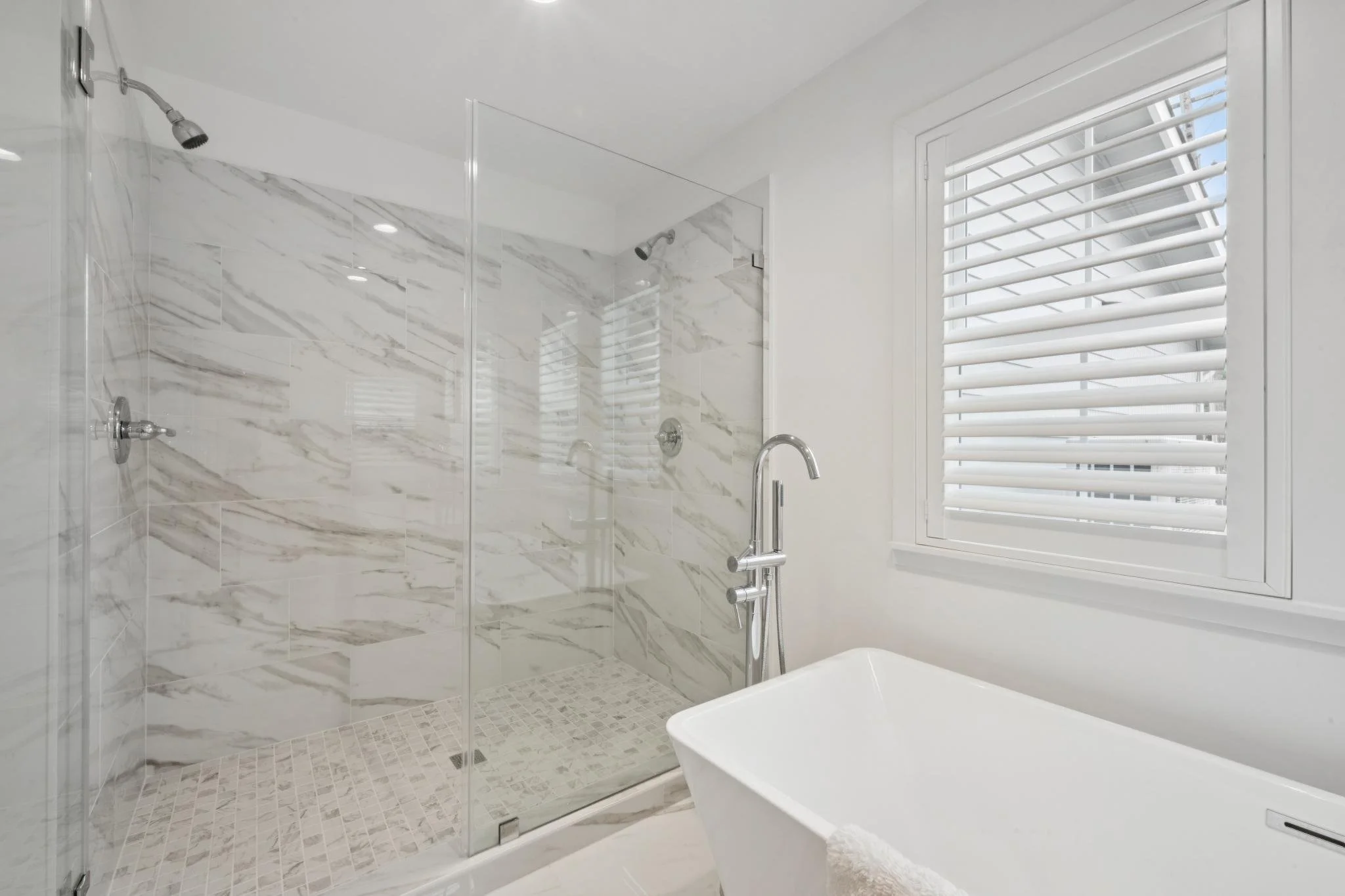 Modern bathroom with a glass-enclosed shower featuring marble tiles, a window with white plantation shutters, and a bathtub with a towel."
