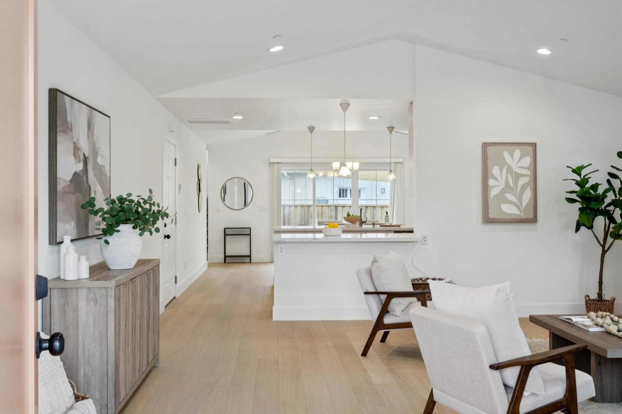 A bright, modern living room and kitchen area with white walls and light wood flooring. Decor includes a large abstract painting, a round mirror, a potted plant, and a wooden console table. A cozy armchair with a white pillow is positioned near a cof