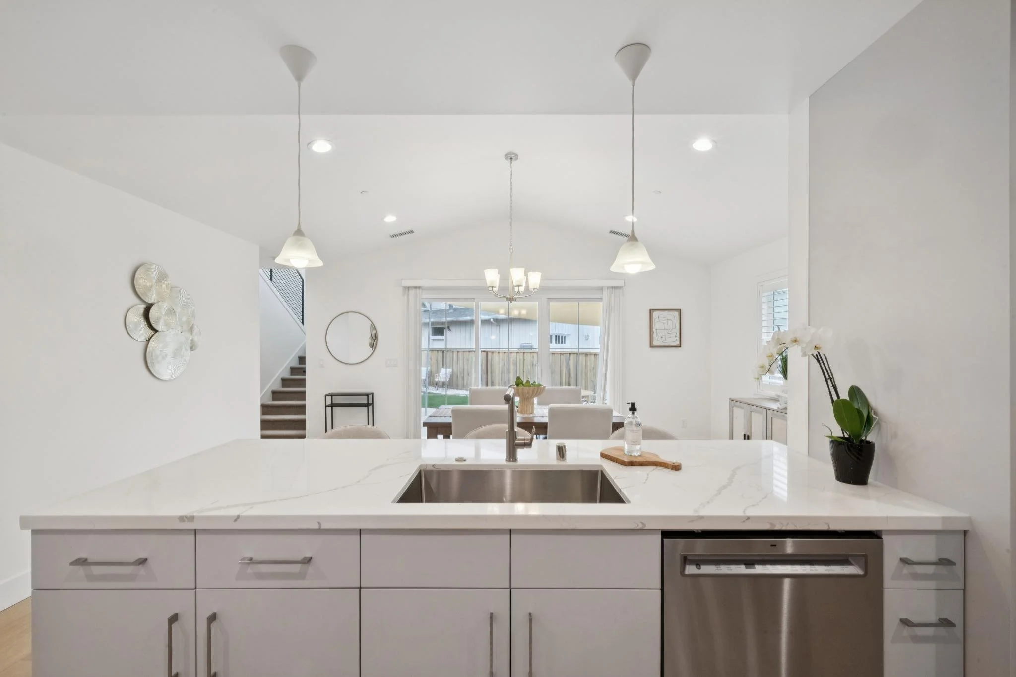 Modern white kitchen with marble countertop island, sink, stainless steel dishwasher, and minimalist decor, including a wall mirror, metal wall art, and potted plant, with a dining area and backyard visible through sliding glass doors.