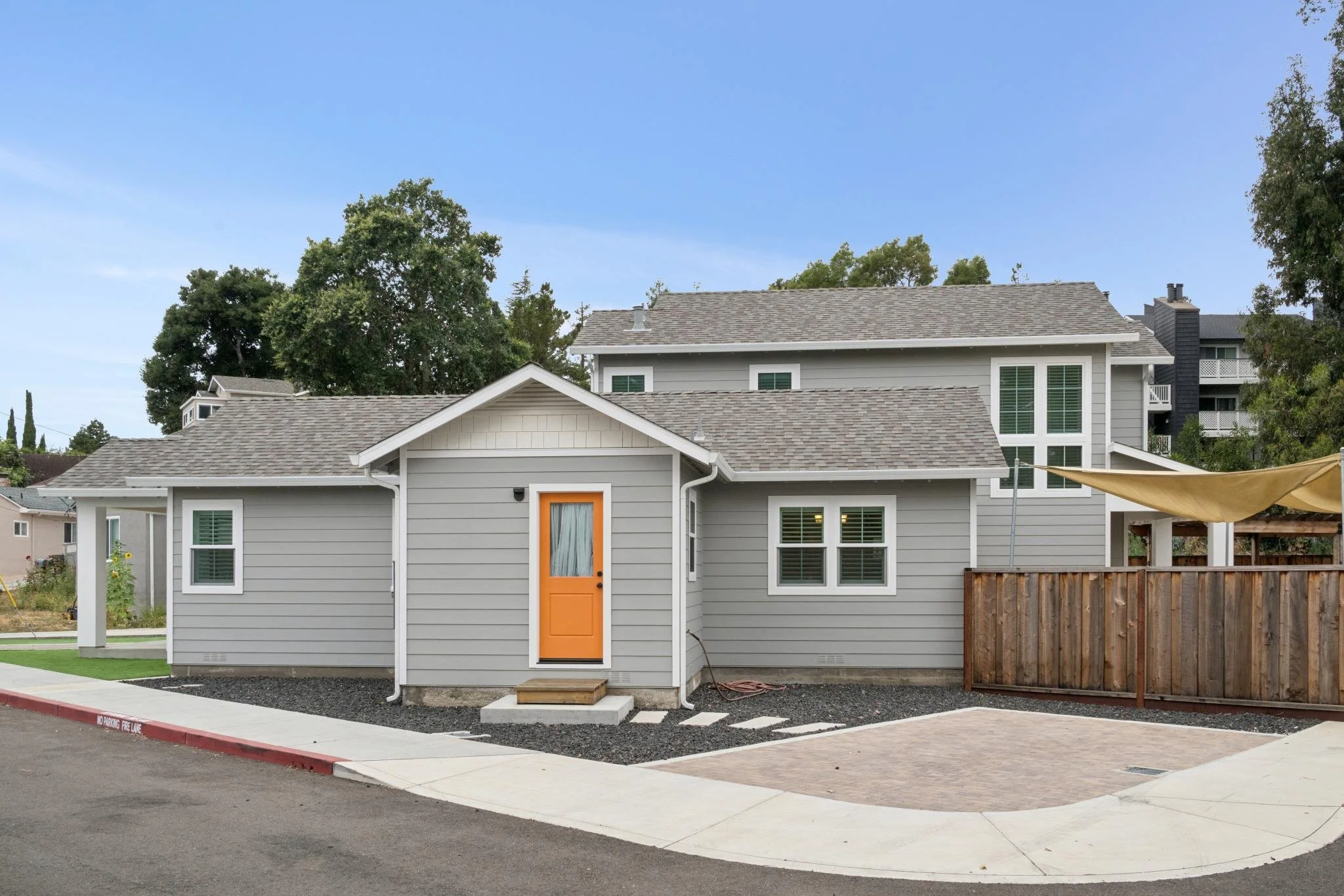 A residential house with gray siding, an orange front door, and windows with shutters. The house has a gabled roof and is situated on a lot with a sidewalk, curb, and a small yard. There are trees and other houses visible in the background.
