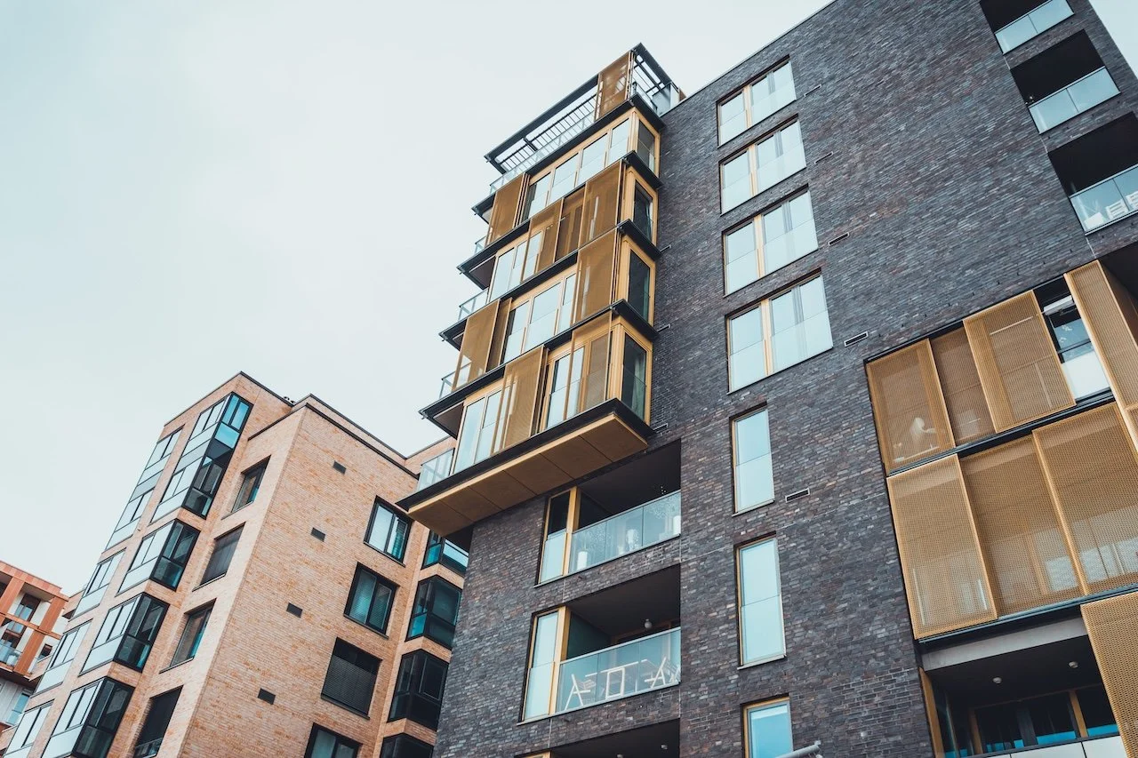 Modern multi-story apartment building with brick and glass exterior, including balconies with yellow and black accents, against a pale sky.