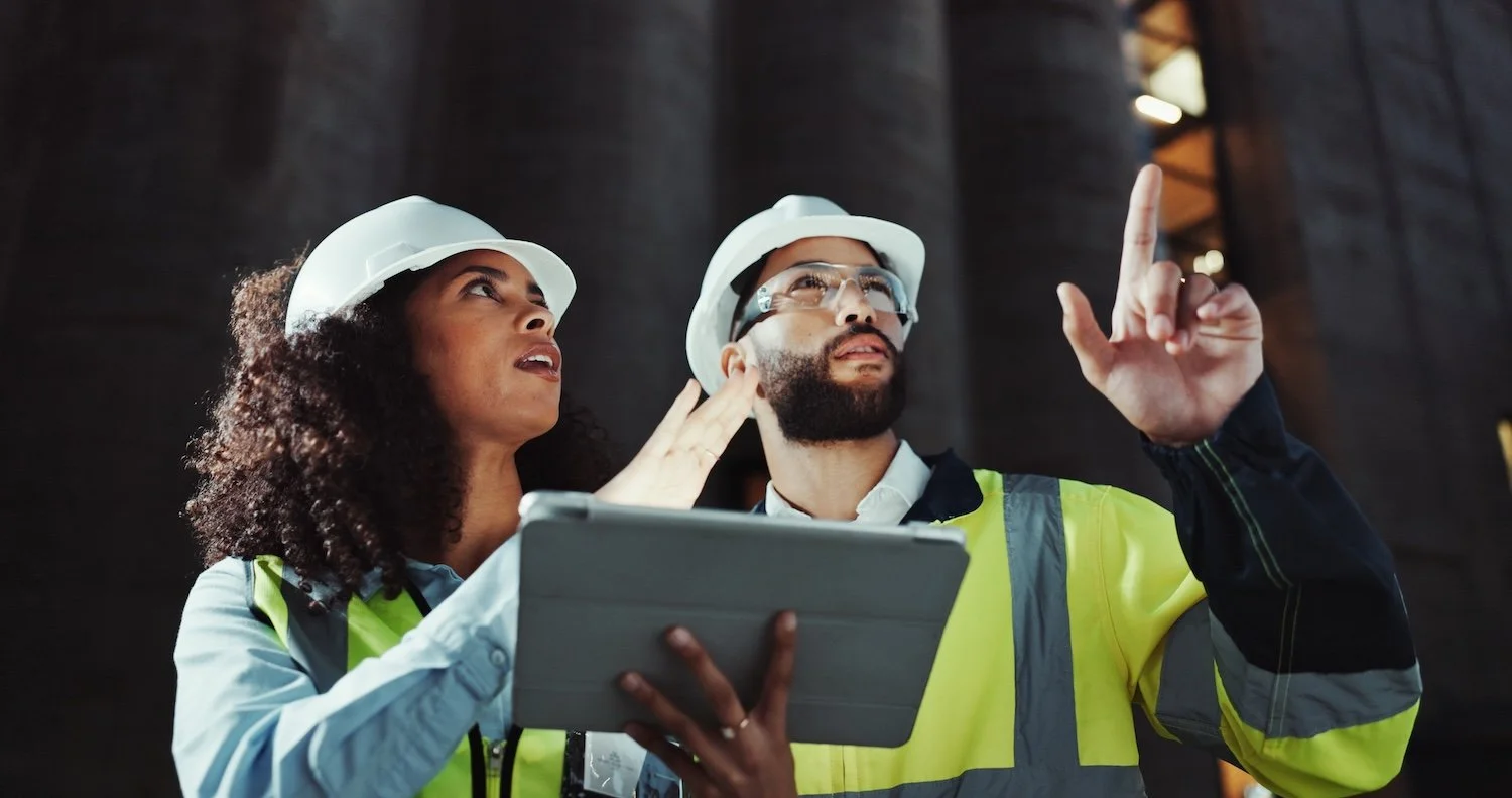 Two construction workers, a woman and a man, wearing safety helmets and high-visibility vests, are inspecting something on-site. The woman is holding a tablet while the man is pointing upward, both looking in the same direction.