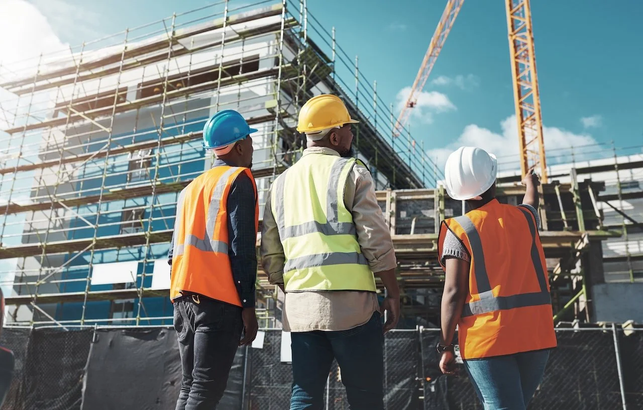Three construction workers, wearing safety helmets and vests, look at a building under construction with scaffolding and a crane in the background.
