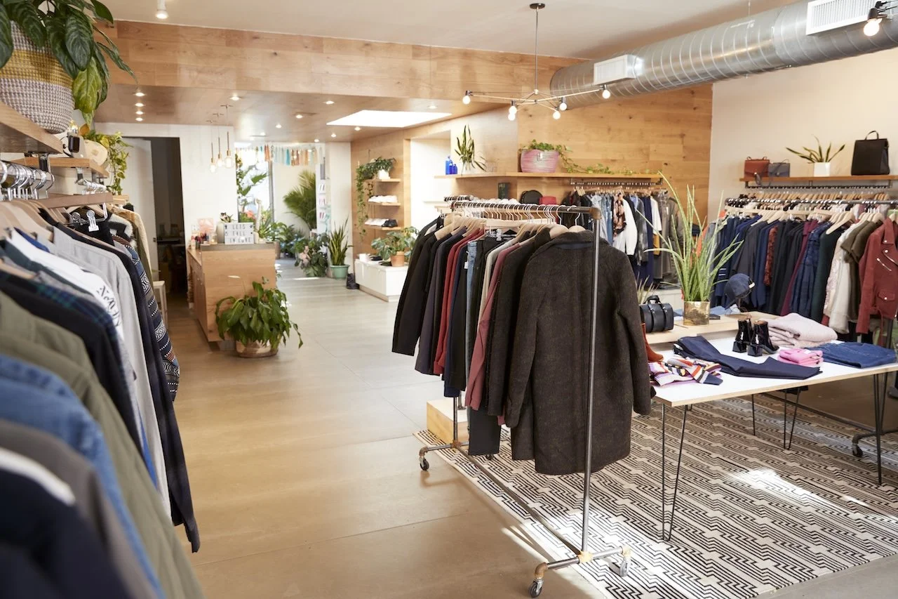 Interior of a clothing store with racks of jackets and shirts, potted plants, a table with folded clothes and accessories, and a wooden decor with warm lighting.