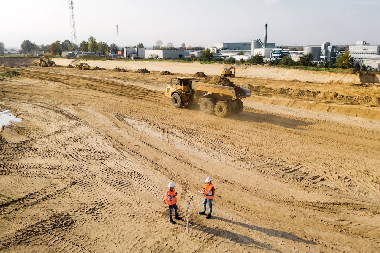 Construction site with large dump trucks moving dirt, two workers with hard hats and safety vests inspecting the site, and industrial buildings in the background.