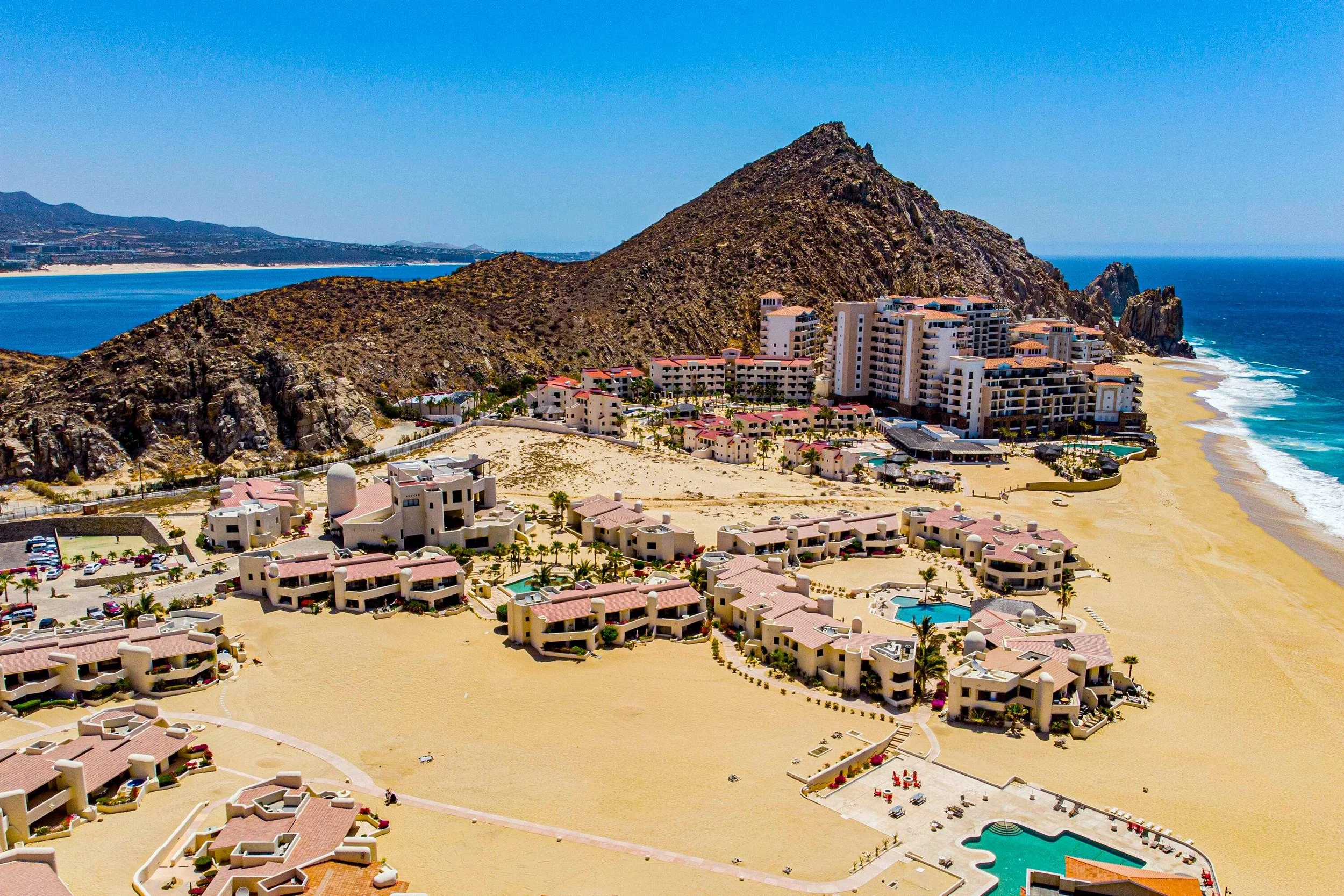 Aerial view of a coastal resort area with multi-story hotels, a beach, rocky hills, and the ocean.