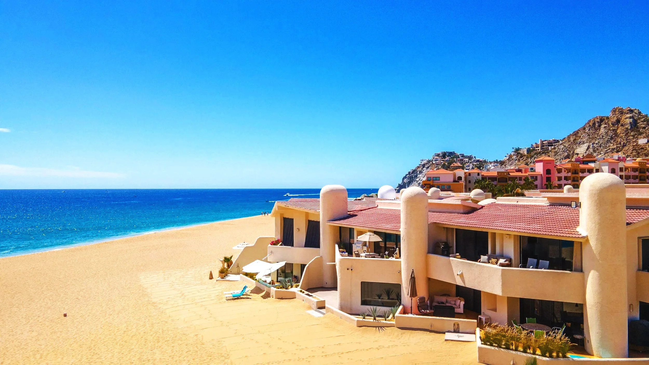 Beachside view of colorful buildings with red rooftops, sandy beach, and blue ocean under a clear blue sky.