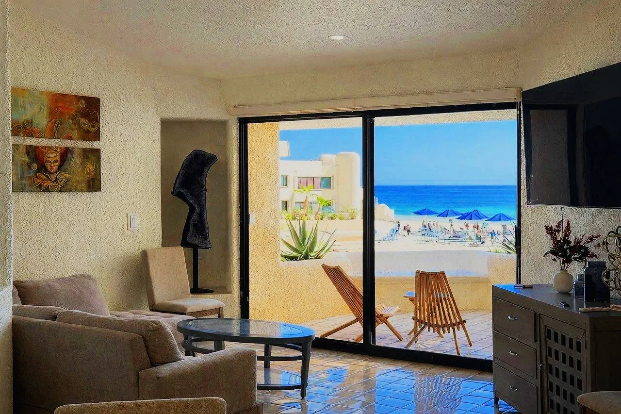 Living room with beige sofa and chairs, art on wall, sliding glass door leading to balcony with wooden chairs, view of beach, umbrellas, and ocean.