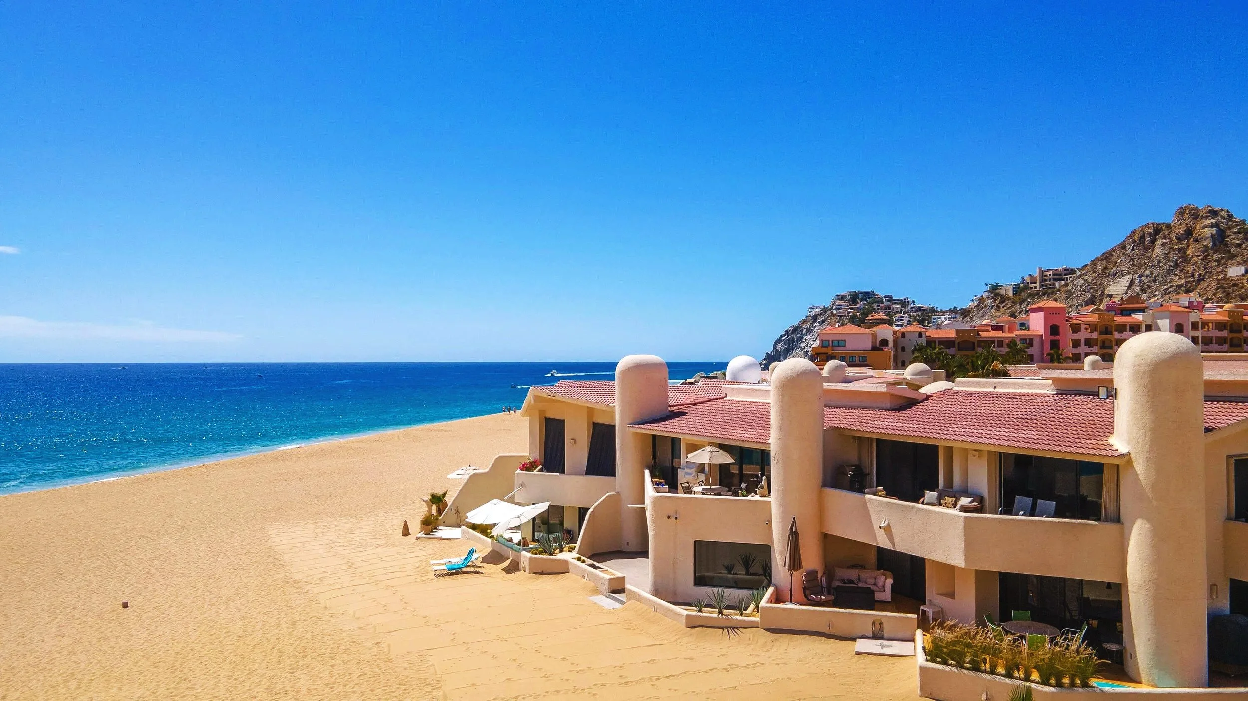 Beachside resort with white buildings, red-tiled roofs, located by sandy beach and blue ocean, with a rocky hillside and pink apartment complex in the background.