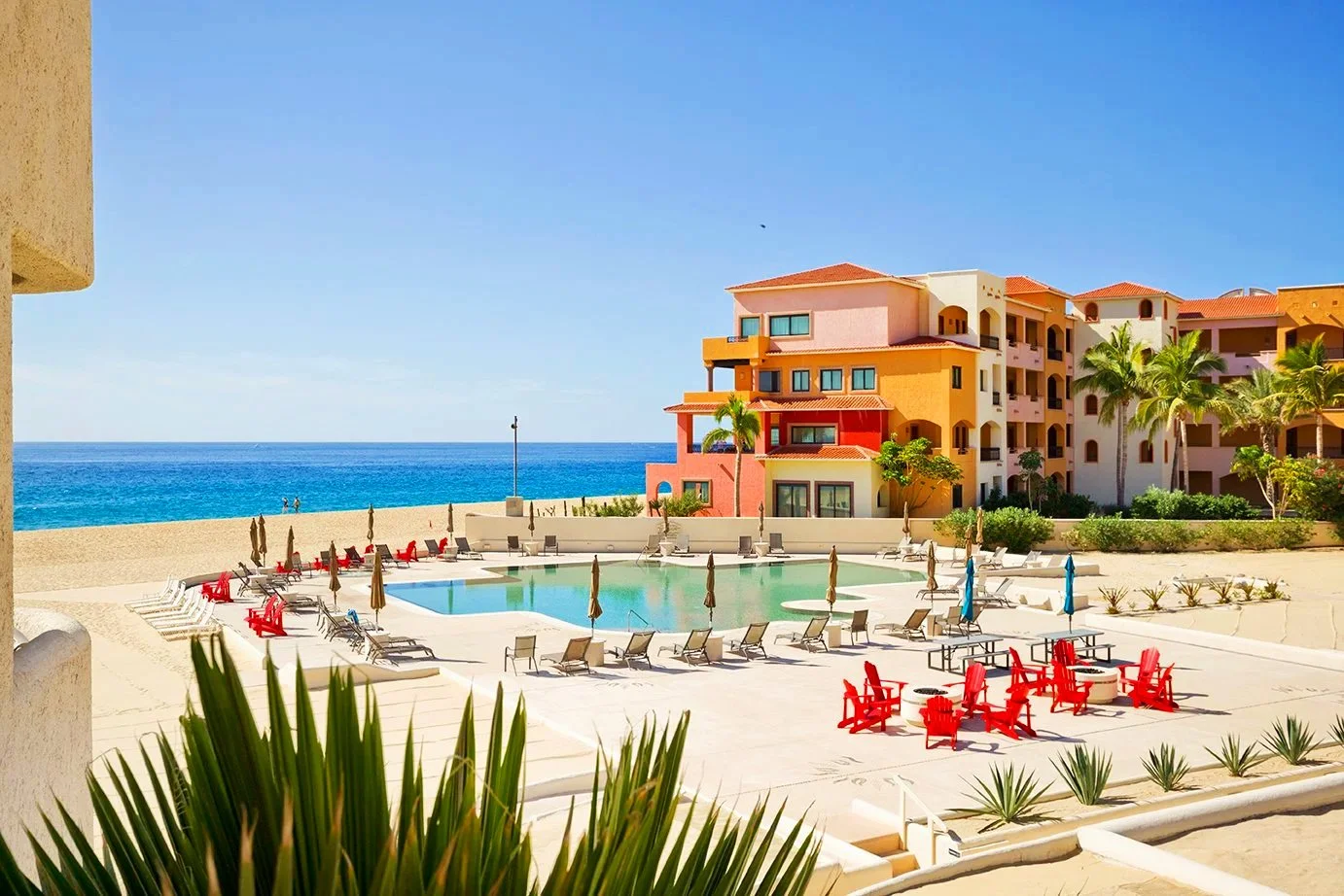 View of a resort pool area with red and beige lounge chairs and umbrellas, surrounded by tropical plants and colorful apartment buildings, with the ocean and a clear blue sky in the background.