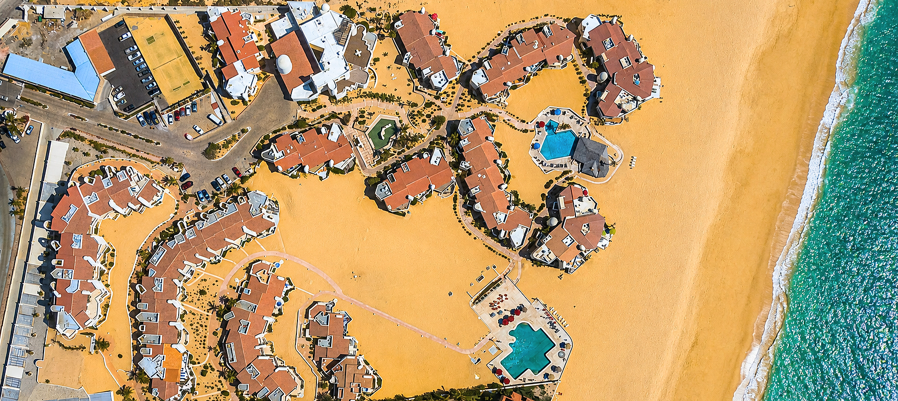 Aerial view of a beachside resort with multiple buildings, swimming pools, and parking lots on sandy terrain next to the ocean.