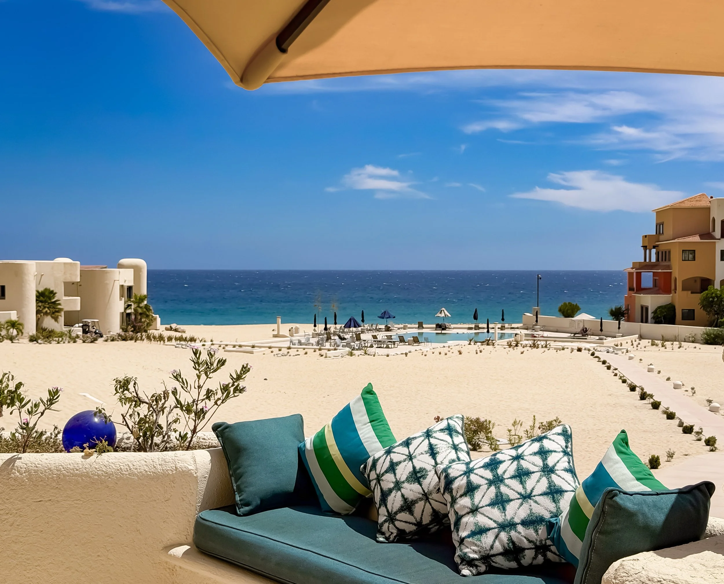 Pillow arrangement on a chair with an ocean view, beige buildings, umbrellas on the beach, blue sky, and a large yellow awning.
